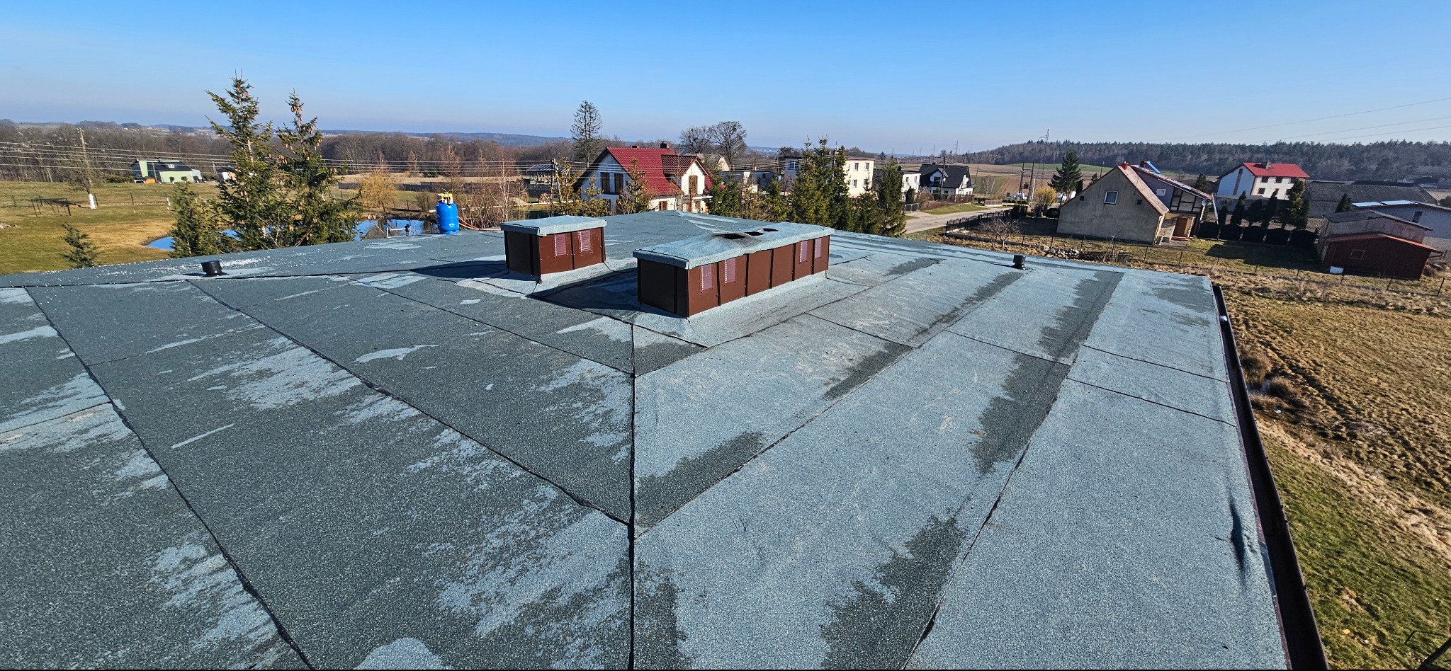 Widok z góry na płaski dach pokryty papą z dwoma brązowymi kominkami wentylacyjnymi. W tle zabudowa wiejska i błękitne niebo.