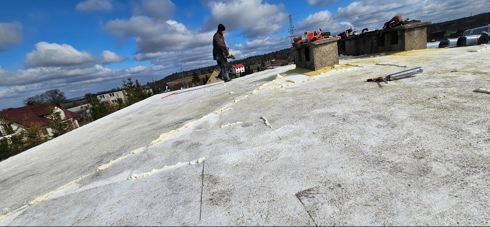 Praca na dachu: dekarz w trakcie uszczelniania połączeń papy termozgrzewalnej pianką montażową. Widoczne narzędzia, komin i fragment zabudowy w tle.