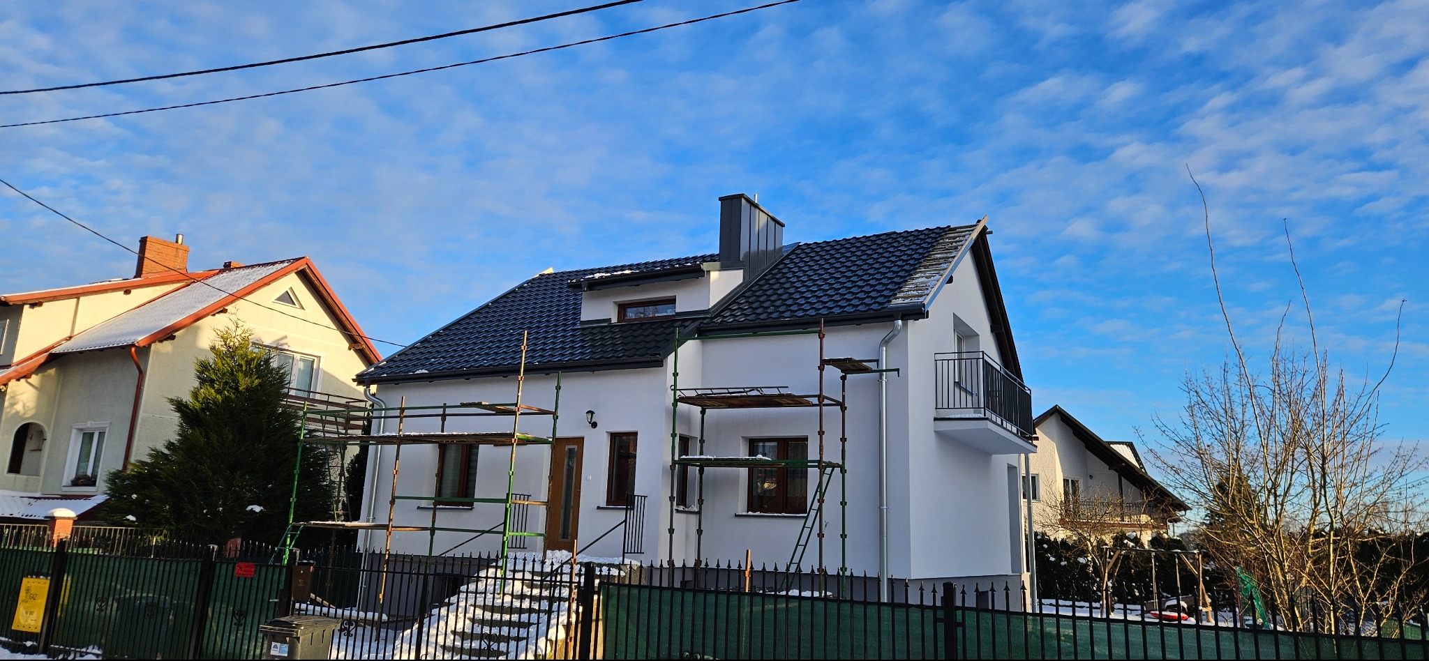 Biały dom jednorodzinny z ciemną dachówką, częściowo otoczony rusztowaniami. Widoczne świeże malowanie elewacji i nowy komin. Błękitne niebo w tle.