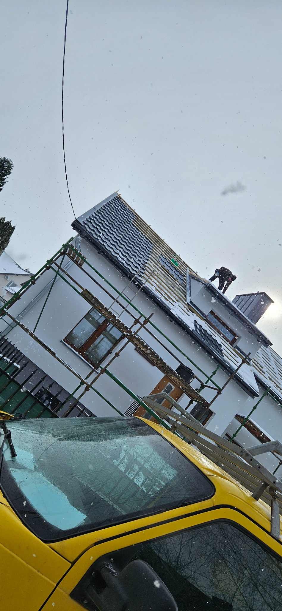 Praca na wysokości: dekarz na rusztowaniu kryje dach domu w zimowej scenerii. Częściowo pokryty dach z ciemnej dachówki, widoczne elementy konstrukcji. Na pierwszym planie żółty samochód.