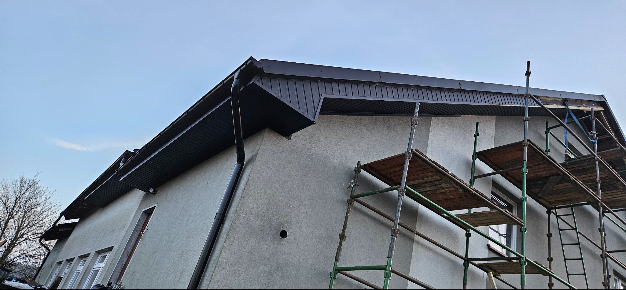 Szarego budynku z czarnym dachem i rynnami, widok z dołu, z rusztowaniem. Dach z blachy trapezowej, elewacja otynkowana. Widoczne okna i fragment drzewa.
