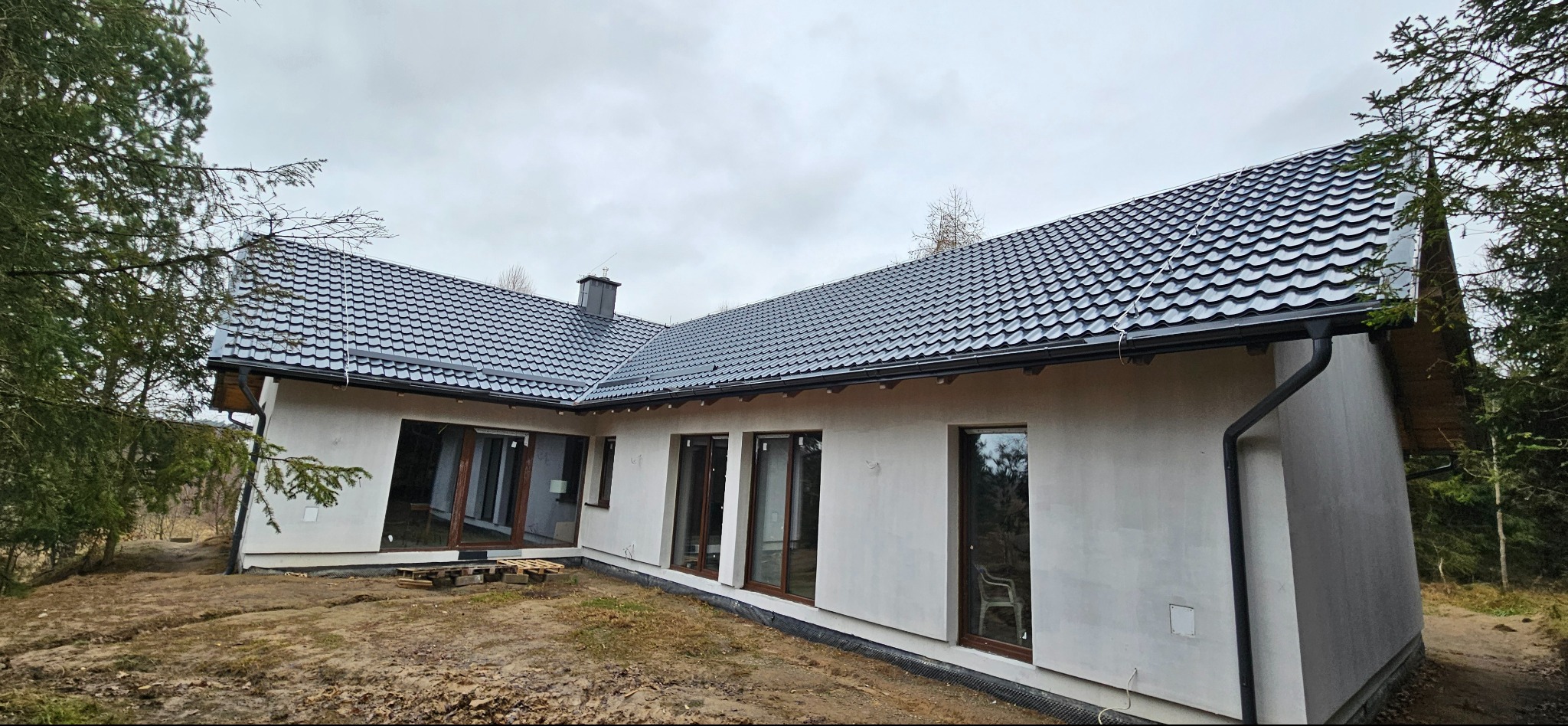 Nowoczesny dom jednorodzinny z ciemną dachówką i rynnami, widoczny z zewnątrz, w trakcie wykańczania elewacji. Otoczenie naturalne, drzewa w tle.
