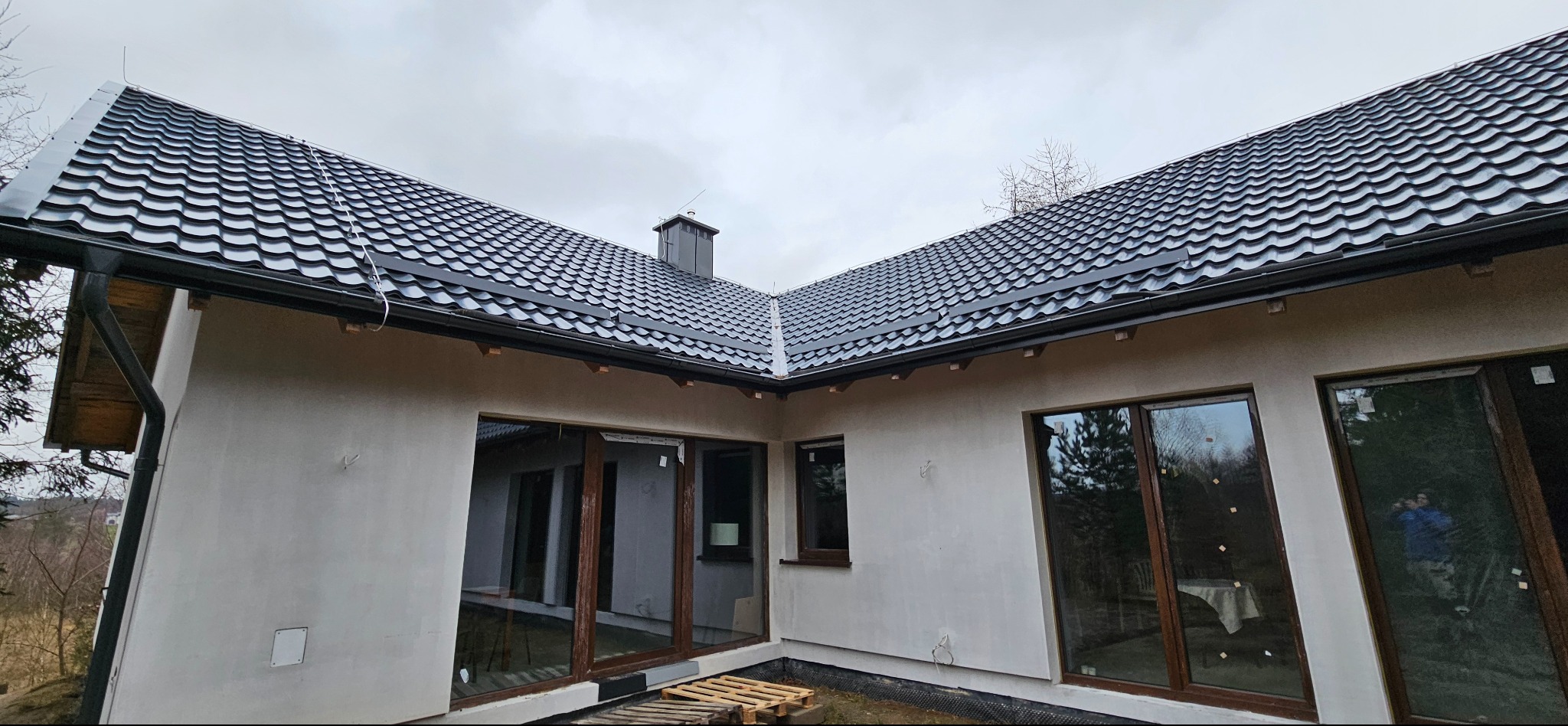 Nowoczesny dom jednorodzinny z ciemną, metalową dachówką i grafitowymi rynnami. Widoczne okna z brązowymi ramami i surowa elewacja. Paleta budowlana na trawniku.