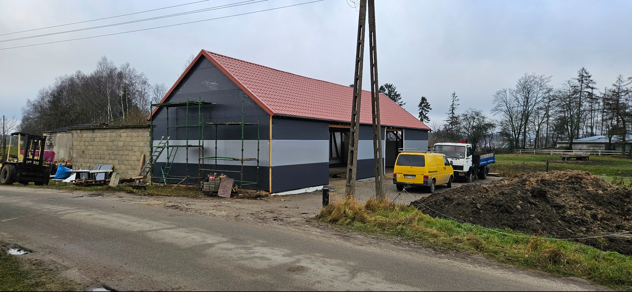 Budynek z nowym, czerwonym dachem i częściowo pomalowaną elewacją w odcieniach szarości. Widoczne rusztowania i pojazdy w tle. Prace w toku.