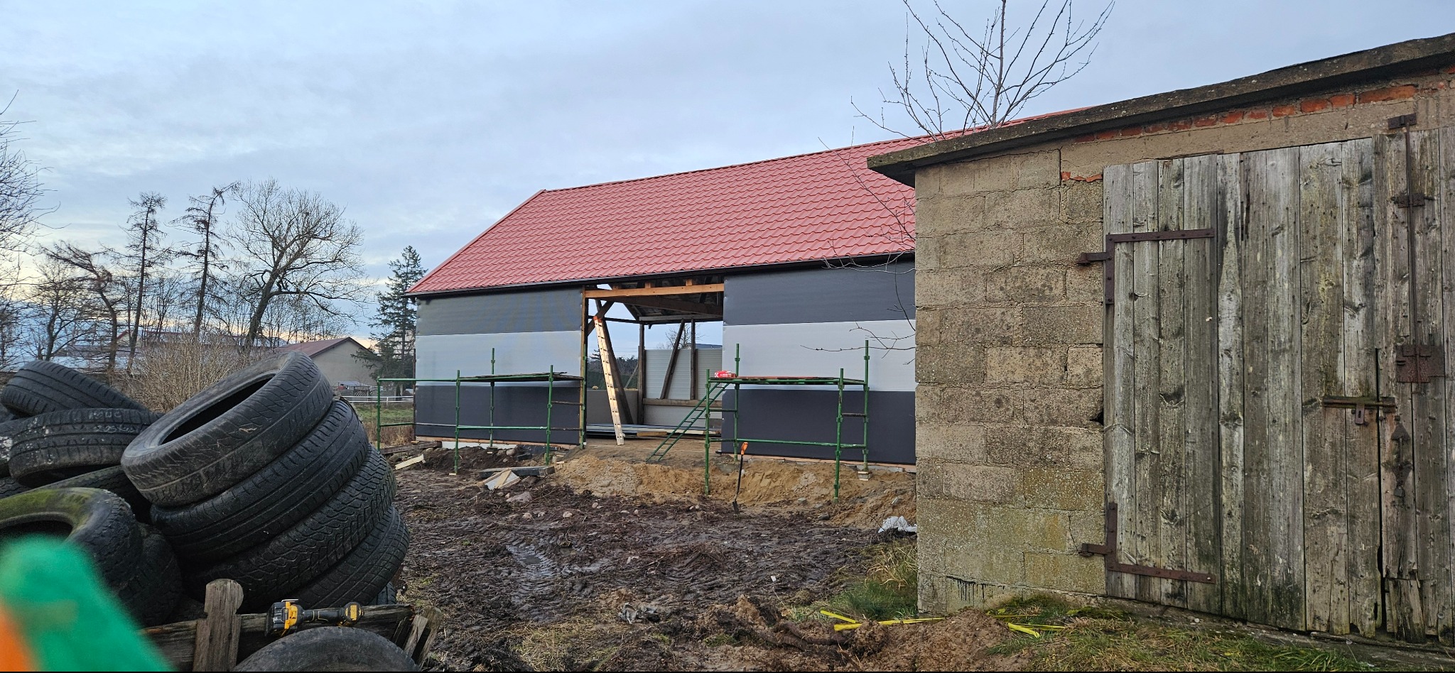 Budynek z czerwonym dachem w trakcie ocieplania elewacji, widoczne rusztowania i przygotowane materiały. Stara stodoła z drewnianymi wrotami w tle.