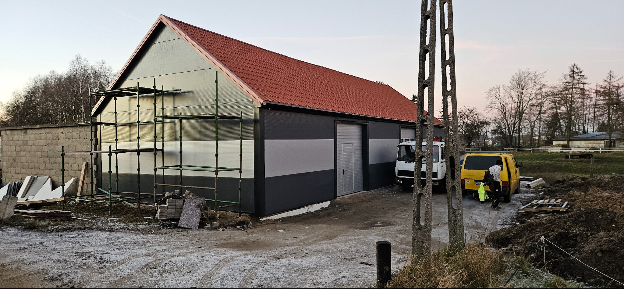 Budynek z nowym, czerwonym dachem i szarą elewacją w trakcie prac wykończeniowych. Widoczne rusztowania, materiały budowlane i pojazdy. Częściowo pochmurne niebo.