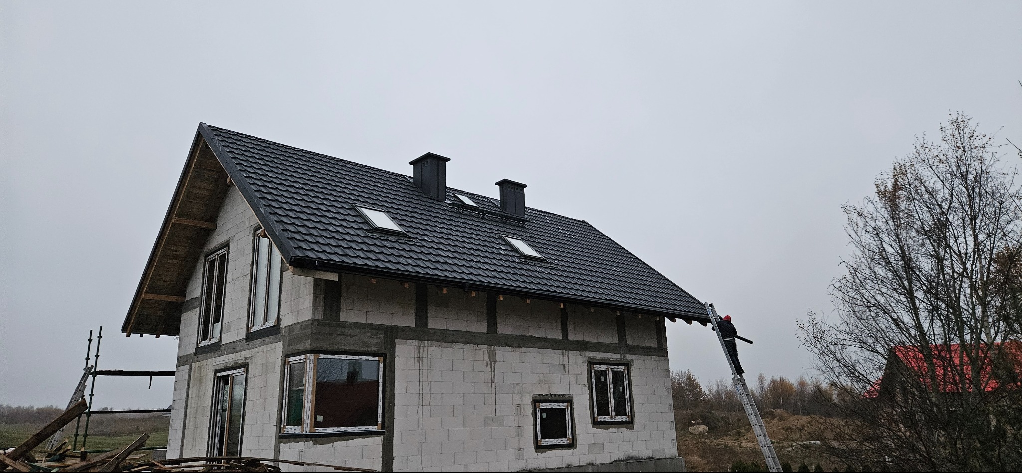 Dom w trakcie budowy z czarnym dachem z dachówki, widoczne okna dachowe i kominy. Pracownik na drabinie montuje rynnę. Surowy stan elewacji.