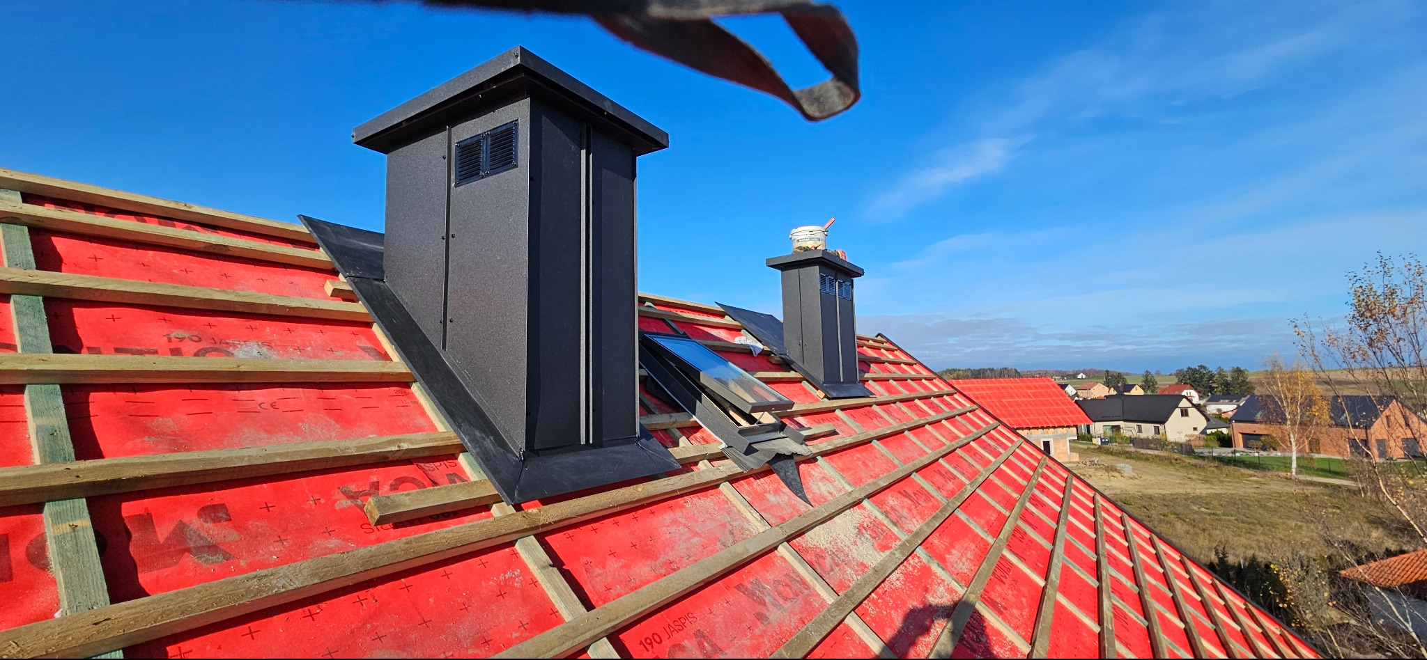 Dach w trakcie budowy z czarnymi kominami i oknem dachowym, widoczne czerwone membrany i drewniana konstrukcja, w tle zabudowa jednorodzinna i błękitne niebo.