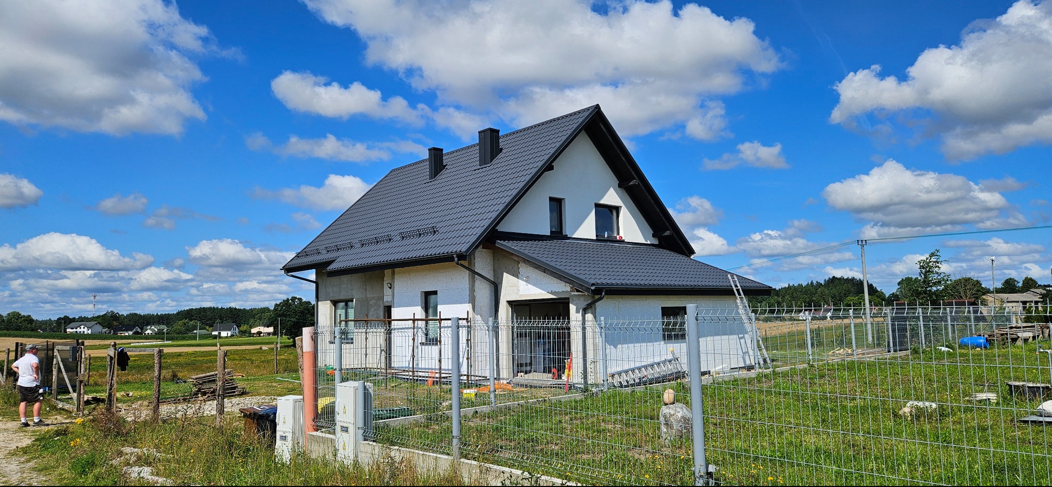 Dom jednorodzinny z ciemnoszarym dachem z blachodachówki, częściowo ocieplony styropianem, ogrodzony siatką. Błękitne niebo z białymi chmurami w tle.