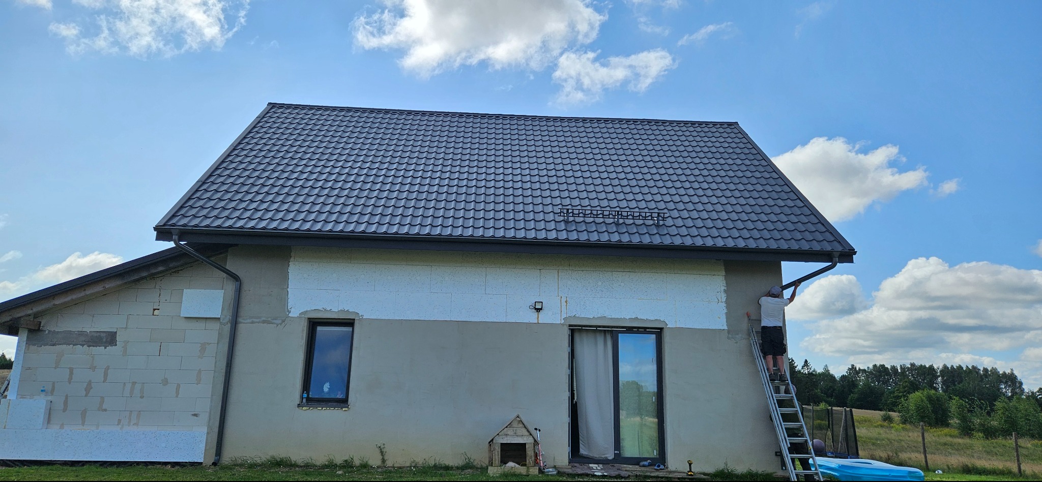 Dom w trakcie ocieplania elewacji z nowym, grafitowym dachem z blachodachówki. Mężczyzna na drabinie montuje rynnę. Widoczne fragmenty ocieplenia styropianem.