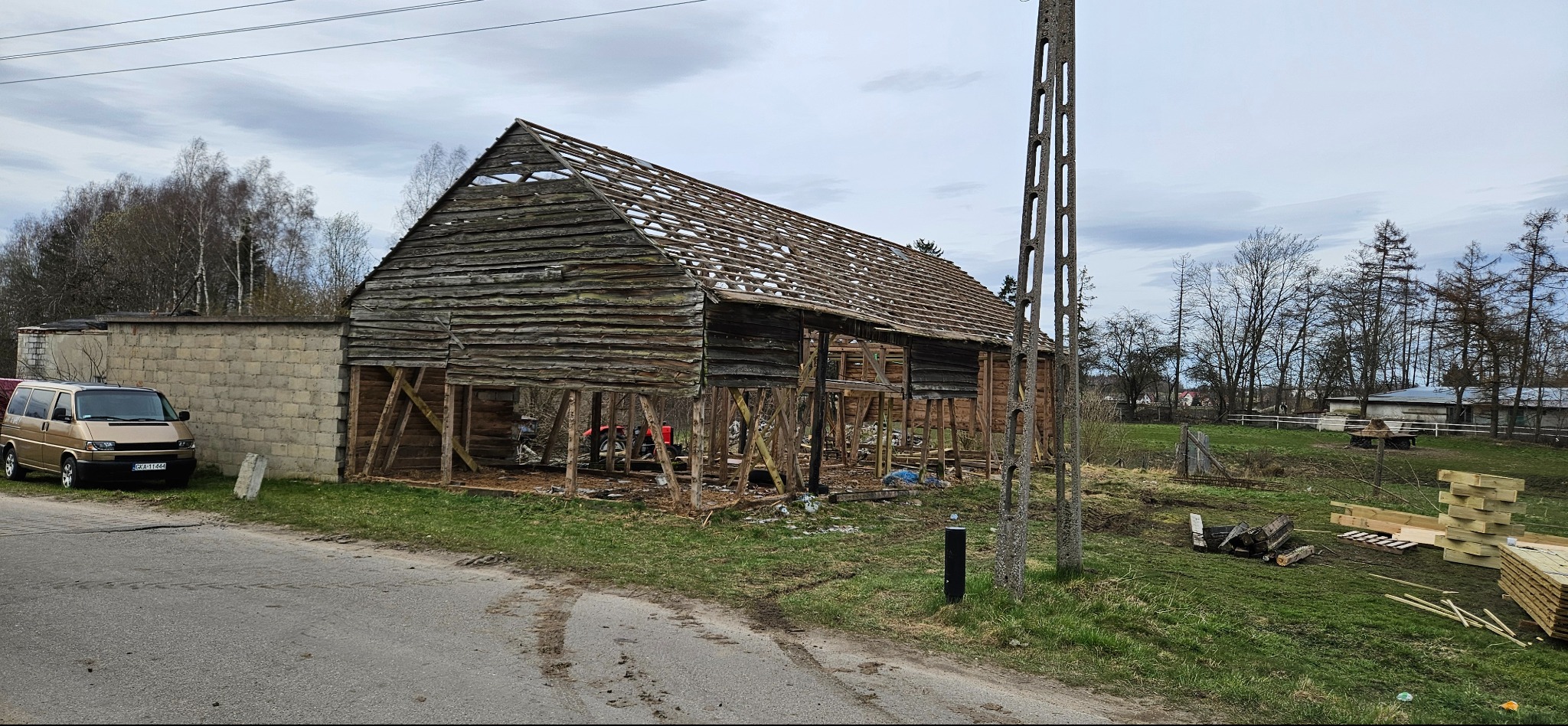 Drewniana stodoła w trakcie remontu dachu, z widocznymi elementami konstrukcyjnymi i stertą nowych desek obok. Na pierwszym planie droga i trawa.