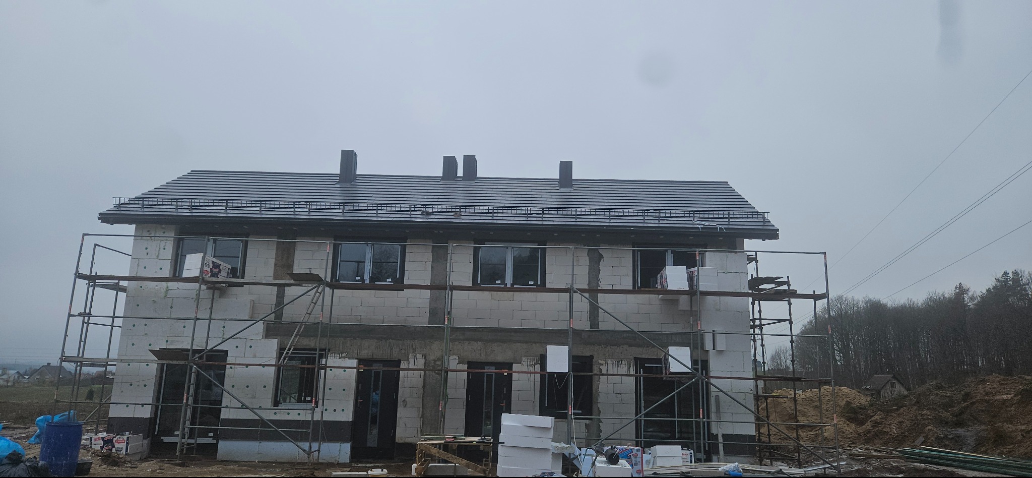 Dwukondygnacyjny dom w trakcie budowy z szarym dachem i rusztowaniami. Widoczne bloczki, okna i materiały izolacyjne. Pochmurne niebo w tle.