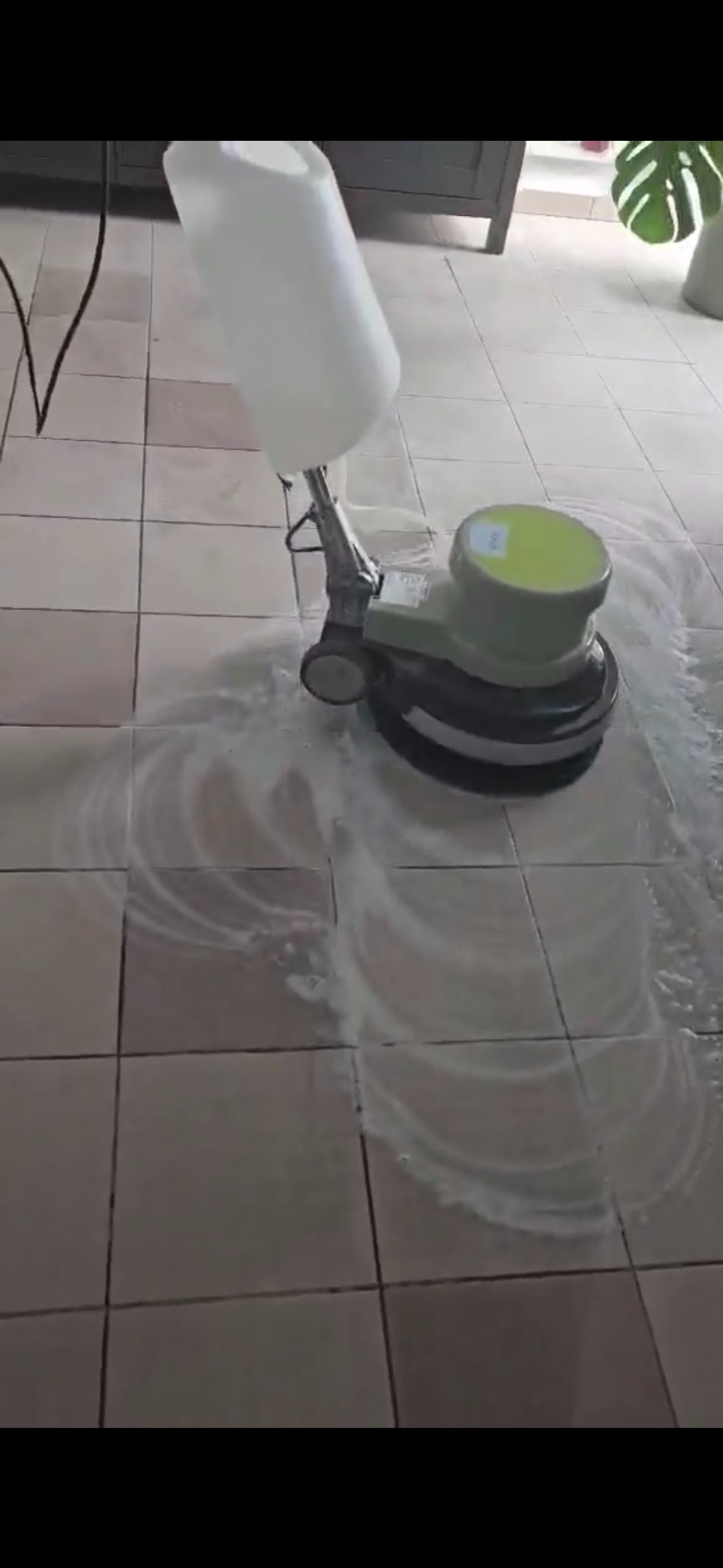 Maszyna do czyszczenia podłóg w akcji, rozprowadza płyn na kafelkach. Widoczny fragment szafki i rośliny doniczkowej w tle. Jasne kafelki poddawane czyszczeniu.