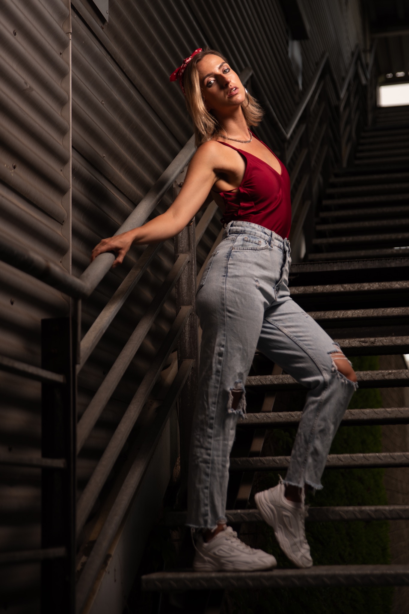 Kobieta w czerwonym topie i jeansach pozuje na metalowych schodach. Ujęcie z dołu, podkreślające sylwetkę. Ciemne tło z blachy falistej i zieleni.