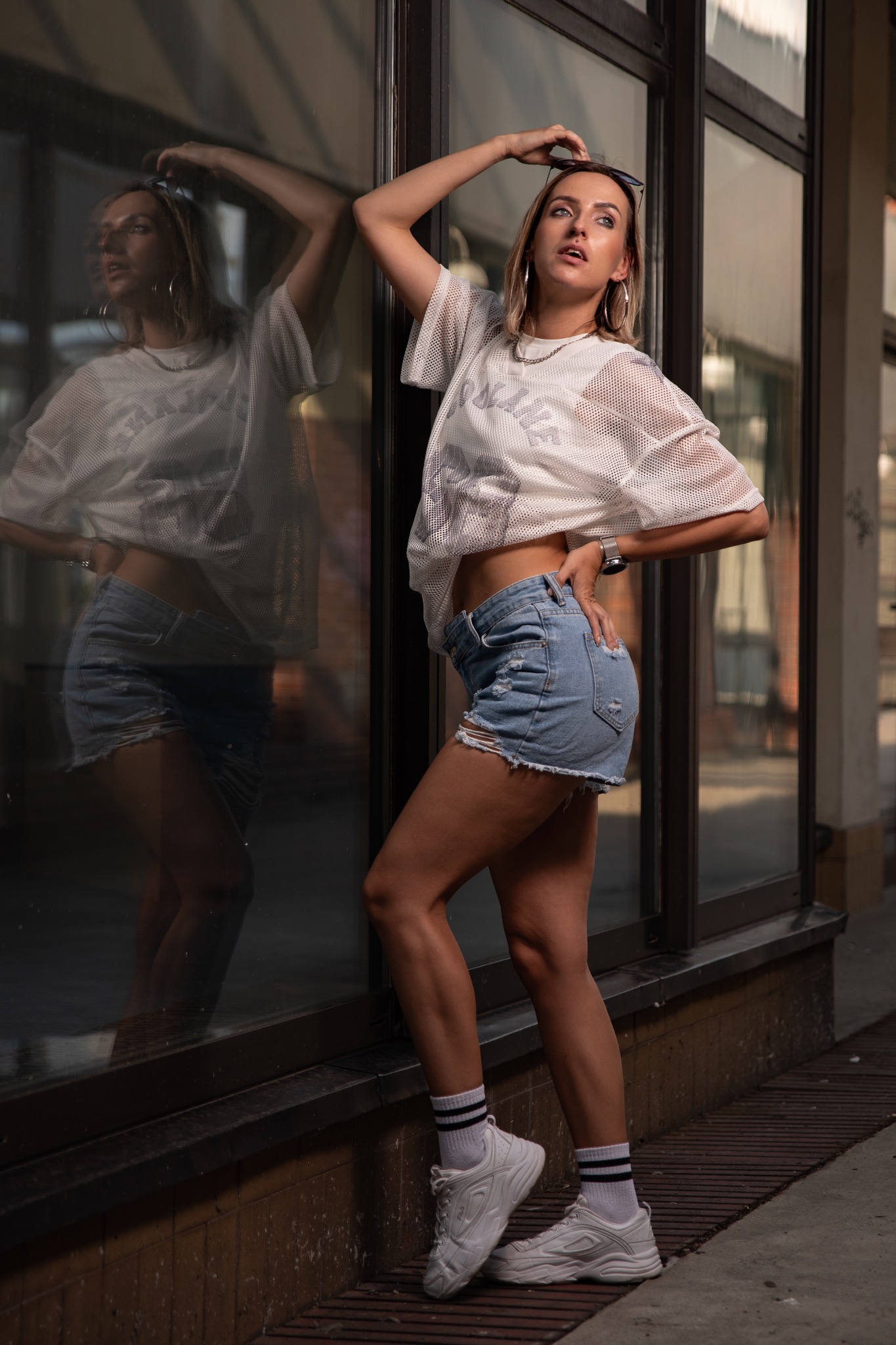 Stylowa blondynka w luźnej koszulce i dżinsowych szortach pozuje przy witrynie sklepowej, odbijając się w szybie. Uwagę zwracają białe sneakersy i skarpetki w paski.