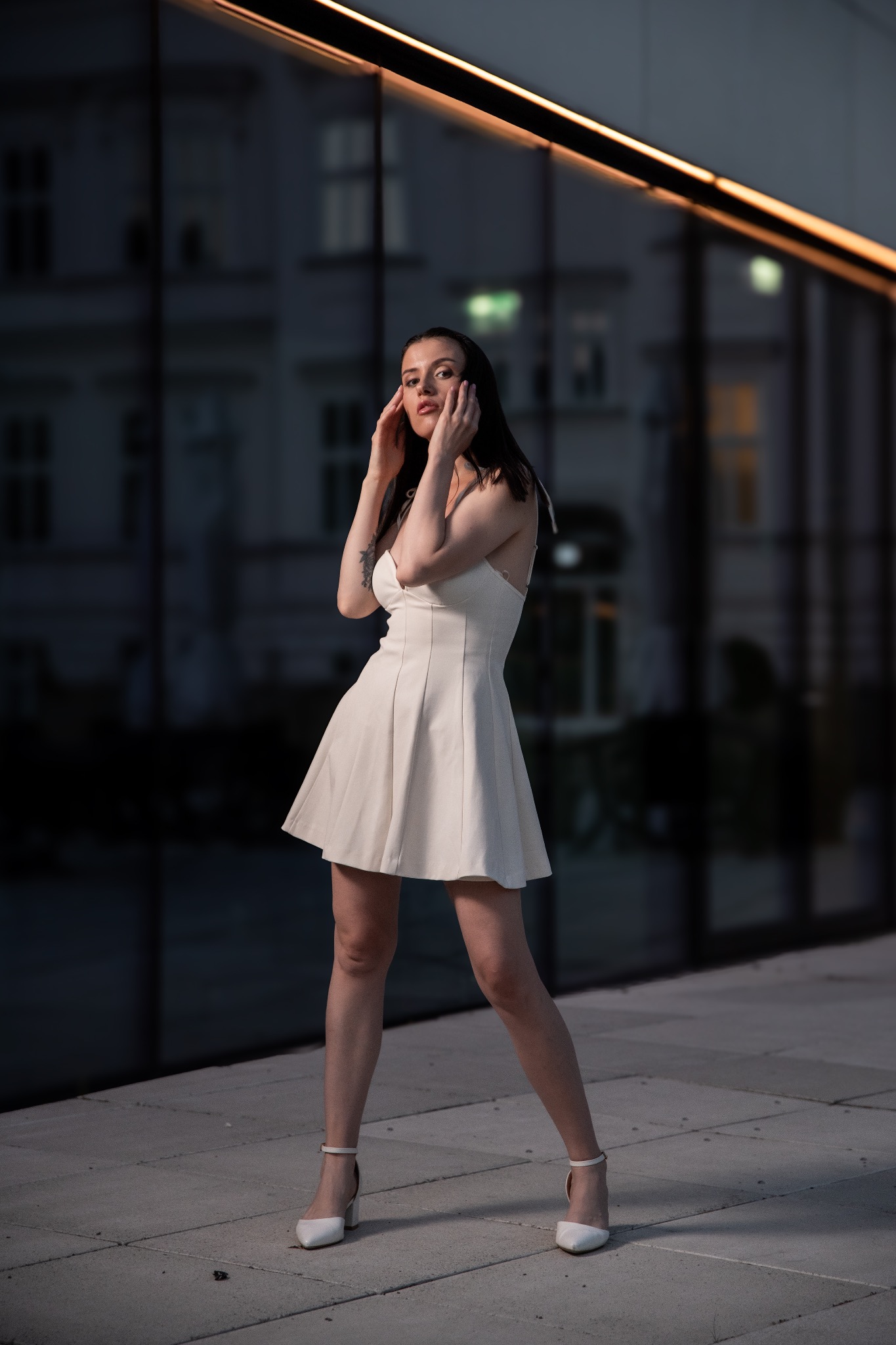 Kobieta w białej sukience pozuje na tle szklanej fasady budynku z odbijającym się światłem. Uwagę zwracają białe buty na obcasie i delikatny makijaż.