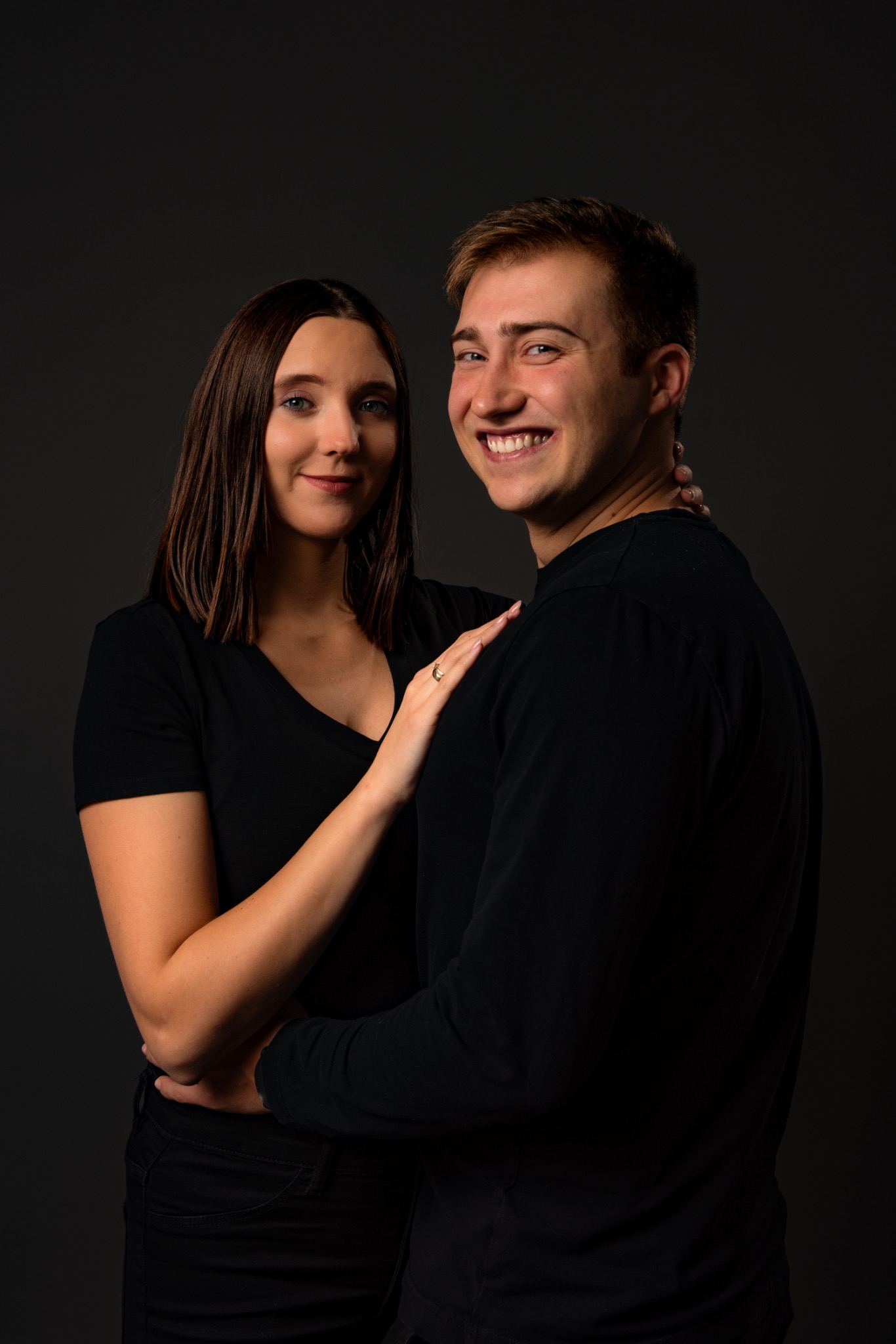 Uśmiechnięta para w czarnych ubraniach pozuje na ciemnoszarym tle. Kobieta z brązowymi włosami i pierścionkiem na palcu obejmuje mężczyznę.