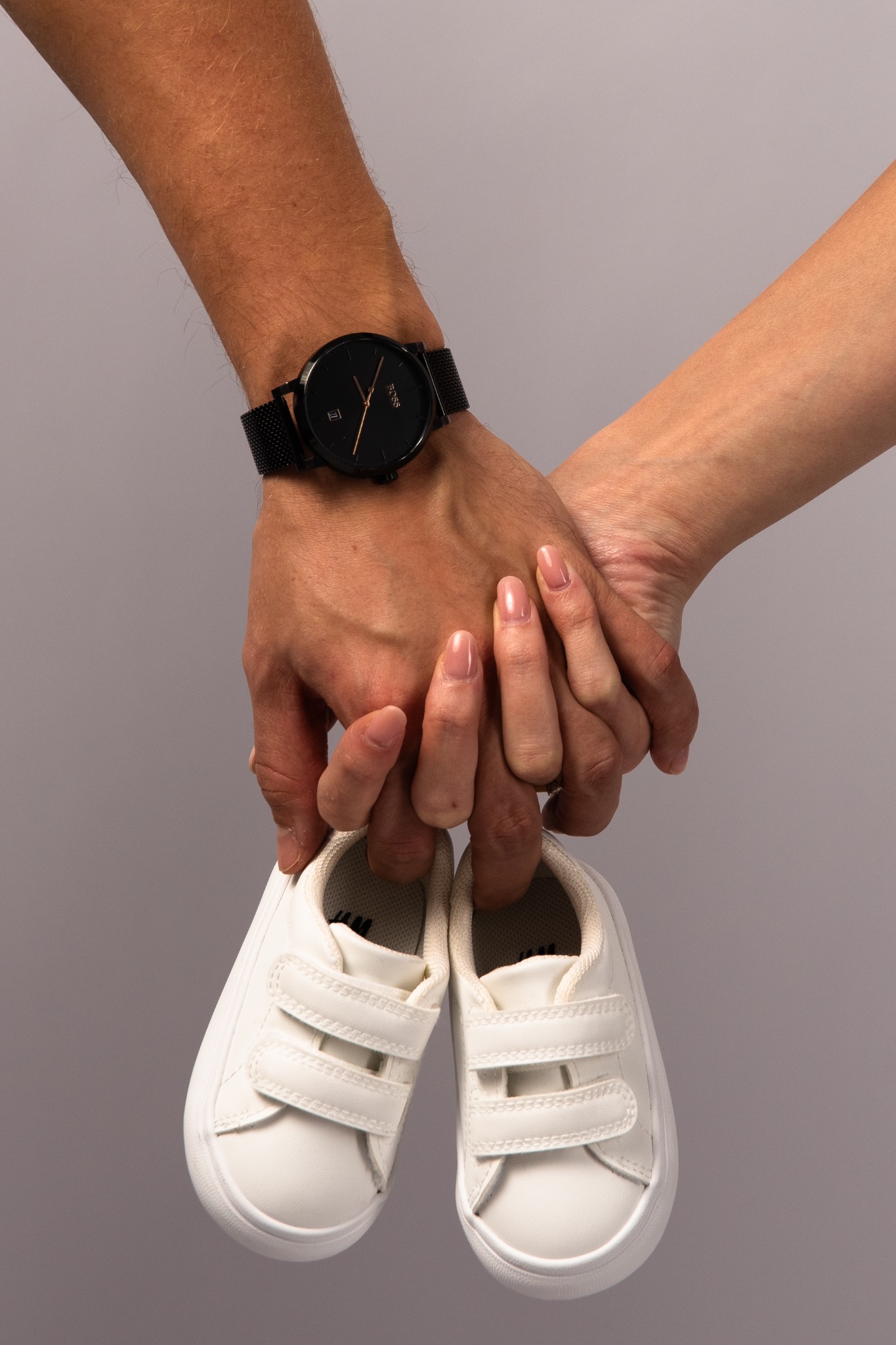 Dłonie kobiety i mężczyzny trzymające białe buciki niemowlęce. Męska dłoń z czarnym zegarkiem. Symbol oczekiwania na dziecko, neutralne tło.