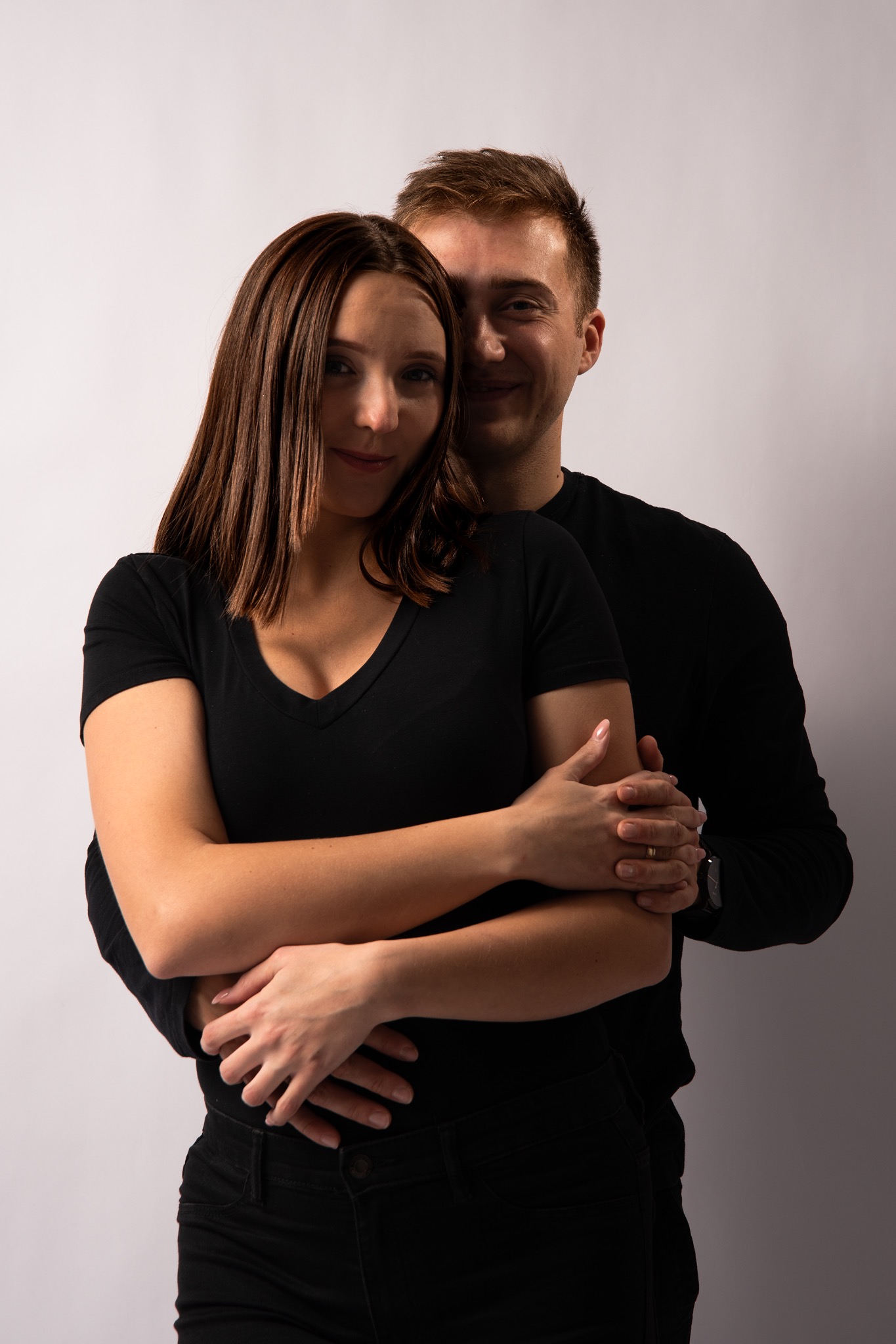 Uśmiechnięta para w czarnych ubraniach, obejmująca się na jasnym tle. Kobieta z brązowymi włosami na pierwszym planie, mężczyzna za nią.