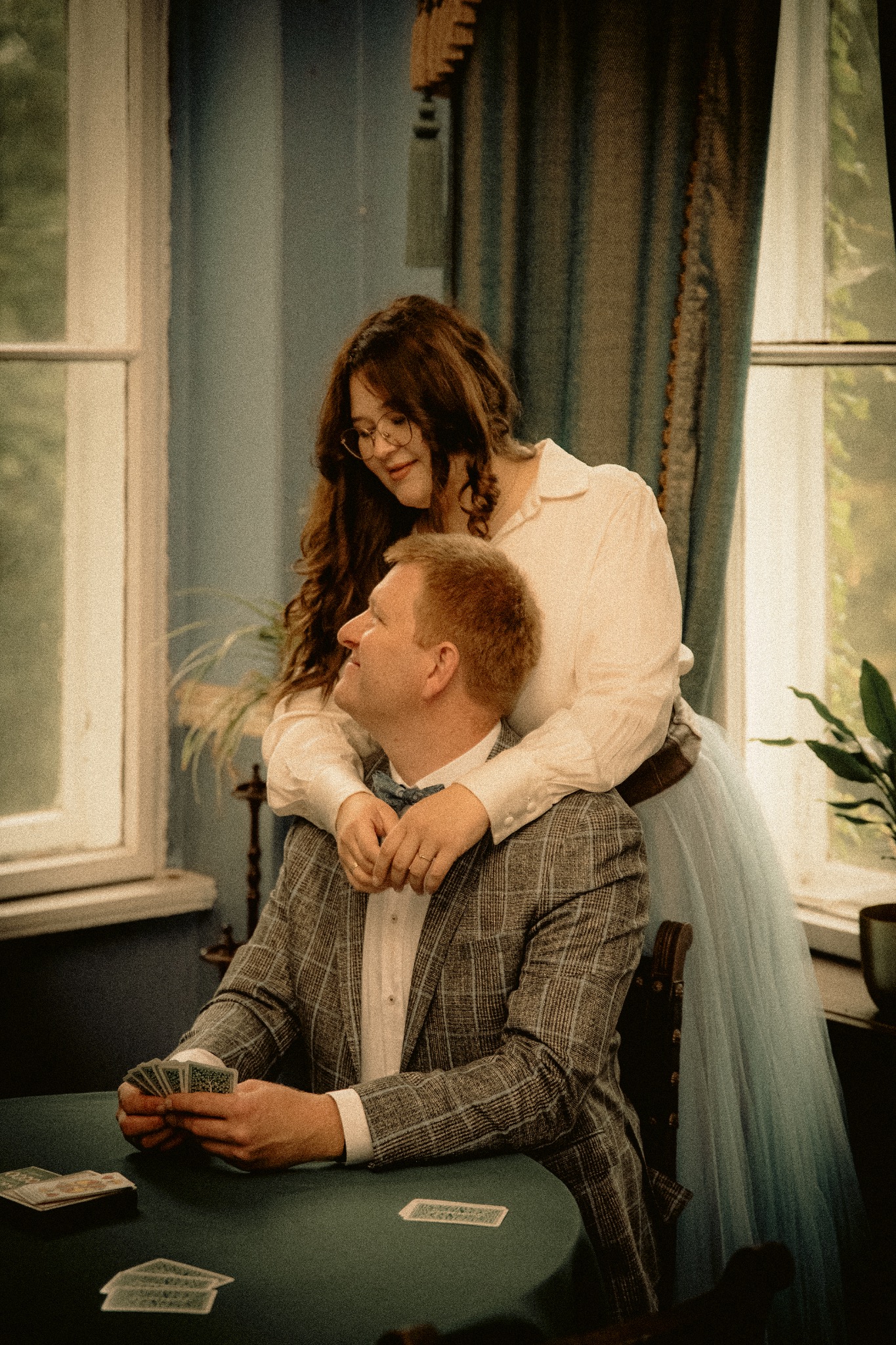 Uśmiechnięta para w stylizowanych strojach: mężczyzna w kraciastej marynarce siedzi przy zielonym stole z kartami, kobieta w okularach obejmuje go od tyłu.