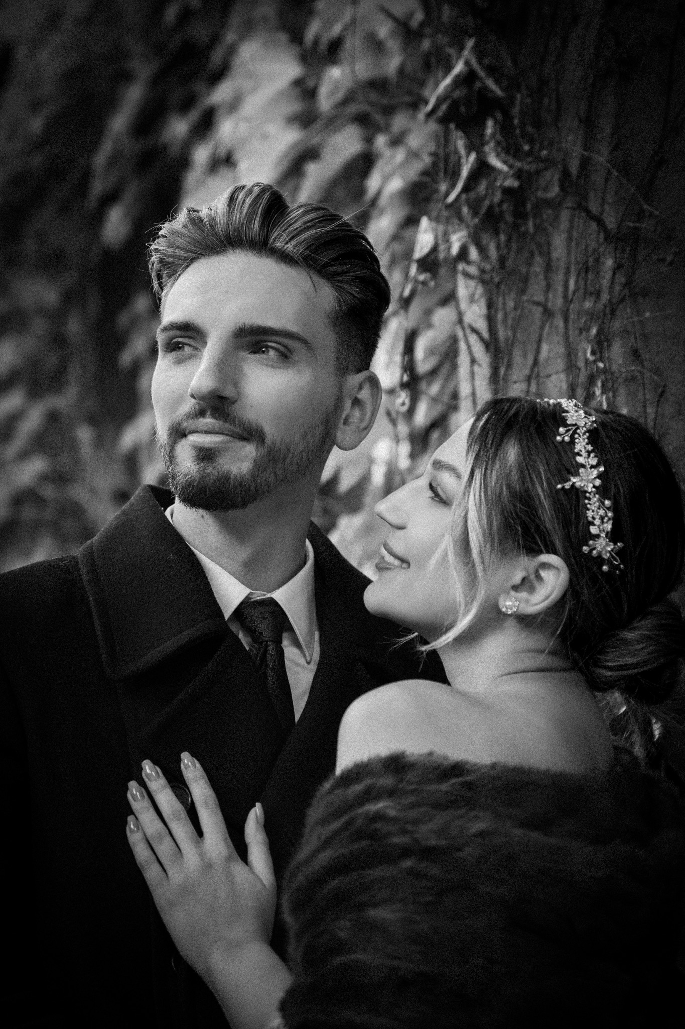 Czarno-białe zdjęcie pary: mężczyzna w płaszczu i kobieta z ozdobą we włosach, obejmująca go. Romantyczny portret w plenerze, z bluszczem w tle.