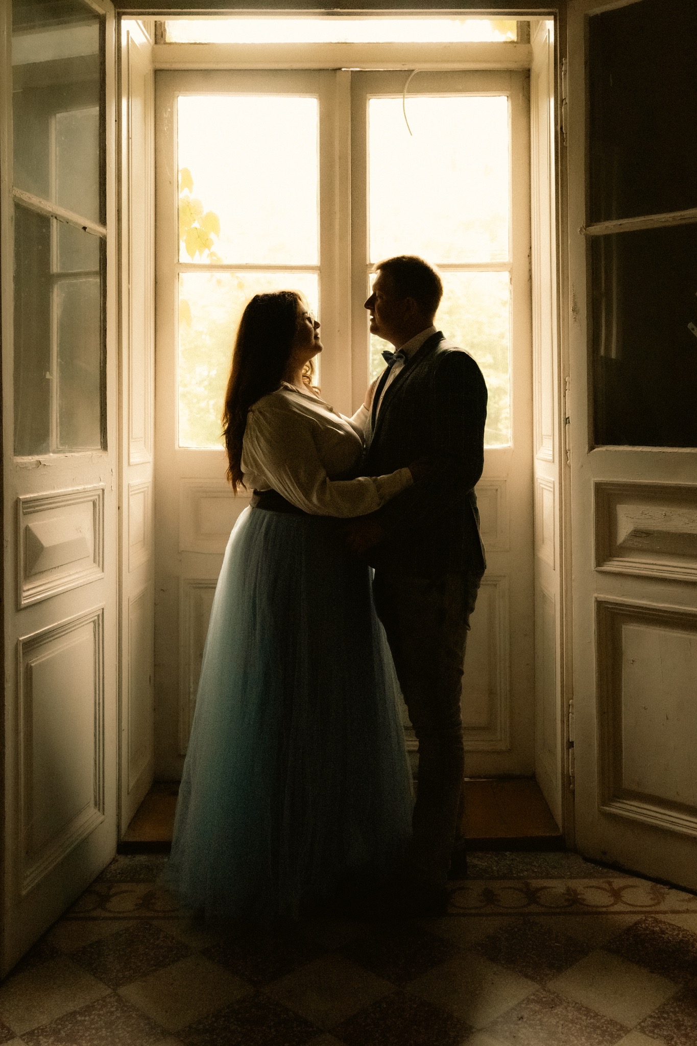 Zakochana para w objęciach na tle okna w starym domu. Kobieta w długiej, błękitnej sukni, mężczyzna w garniturze z muchą. Romantyczna sesja w klimatycznym wnętrzu.