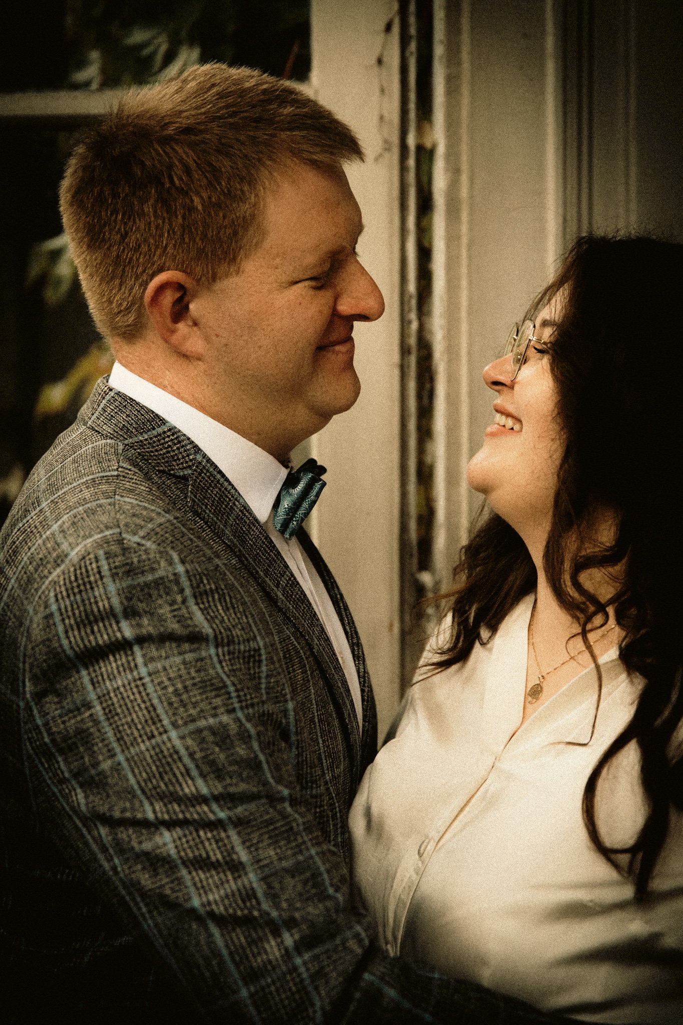 Uśmiechnięta para w zbliżeniu: mężczyzna w kraciastej marynarce i muszce, kobieta w okularach i bluzce. Romantyczna sesja w ciepłym oświetleniu.