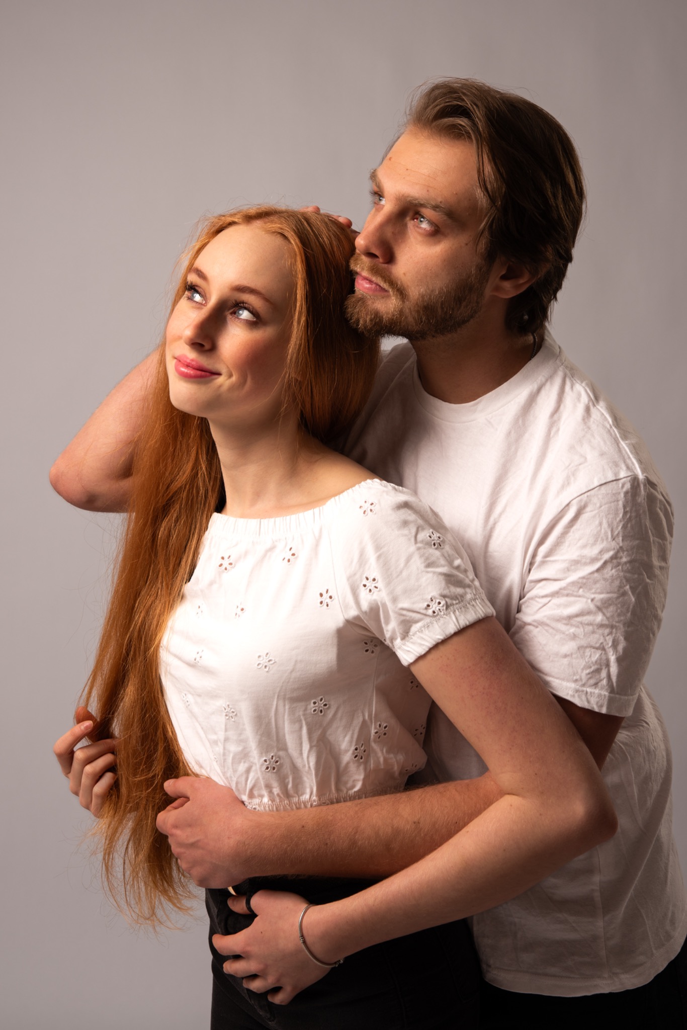 Para w objęciach, ona z rudymi włosami w białej bluzce, on z brodą w białym t-shircie, oboje patrzą w górę, na jasnoszarym tle. Romantyczna sesja.