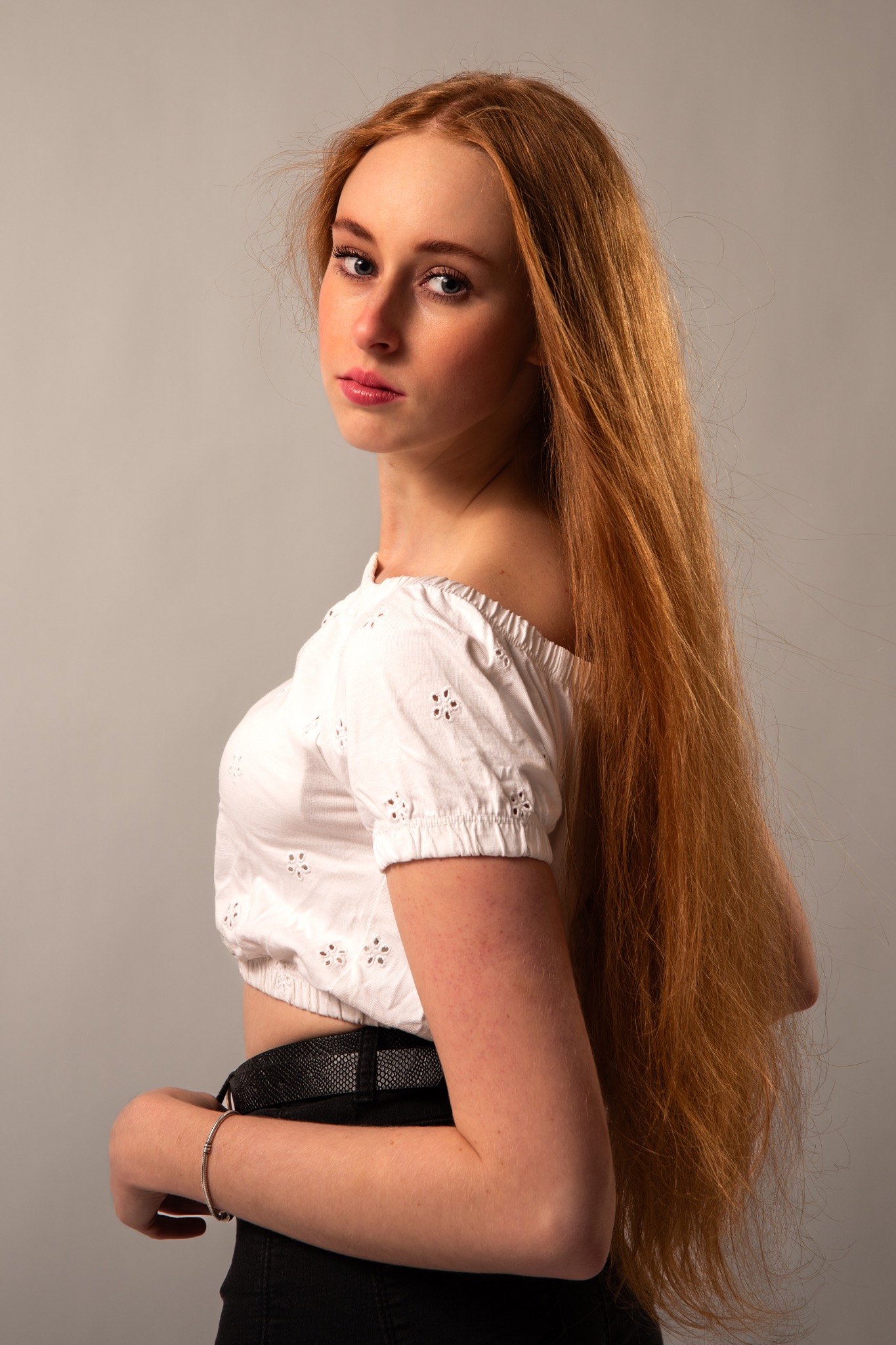 Portret młodej kobiety z długimi, rudymi włosami, ubrana w białą bluzkę z haftem, pozująca na szarym tle. Ujęcie z profilu, z delikatnym spojrzeniem w stronę obiektywu.