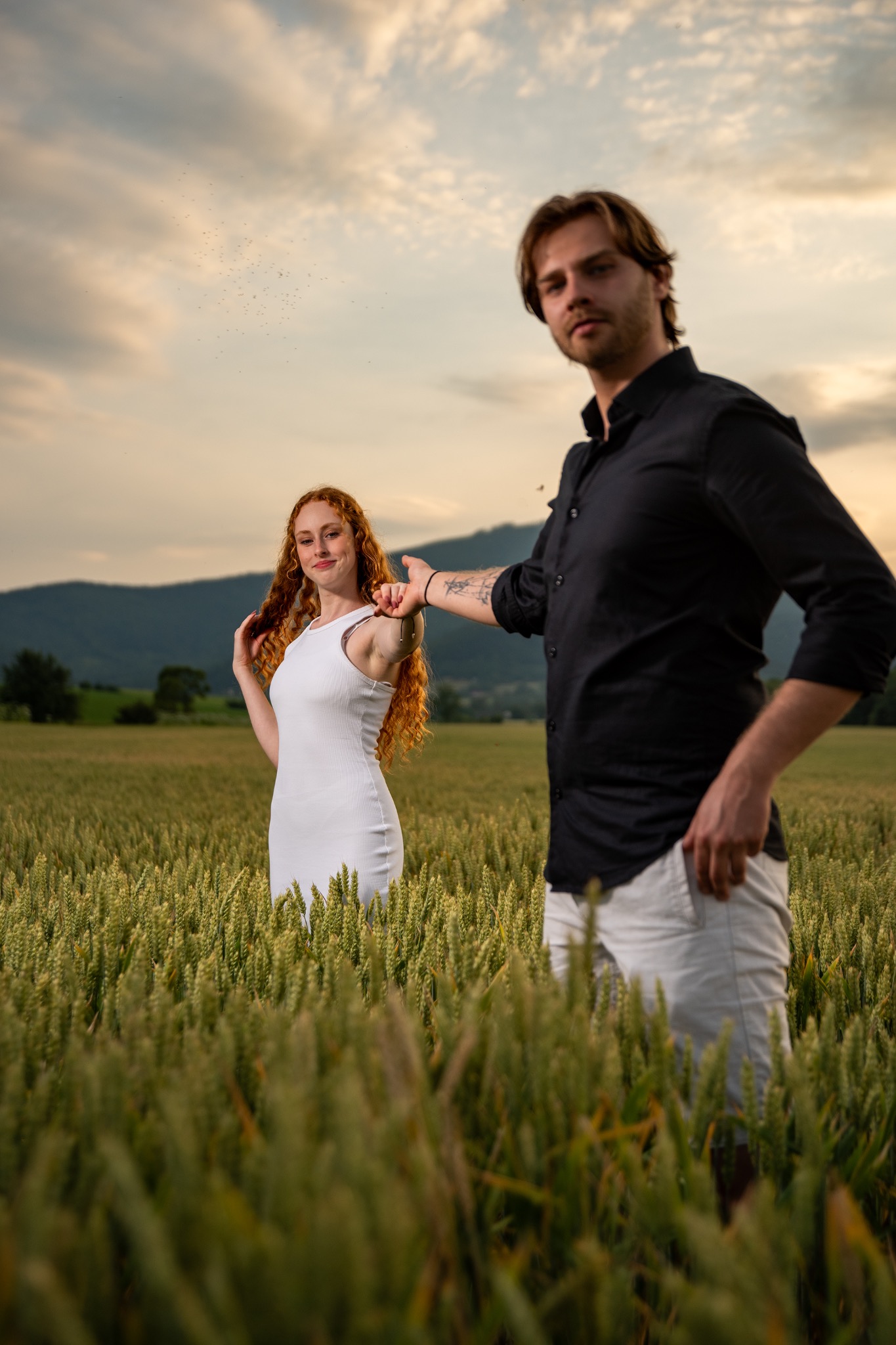 Para w polu zboża podczas sesji narzeczeńskiej. Kobieta w białej sukience, mężczyzna w czarnej koszuli. Romantyczna sceneria z górami w tle i zachodzącym słońcem.
