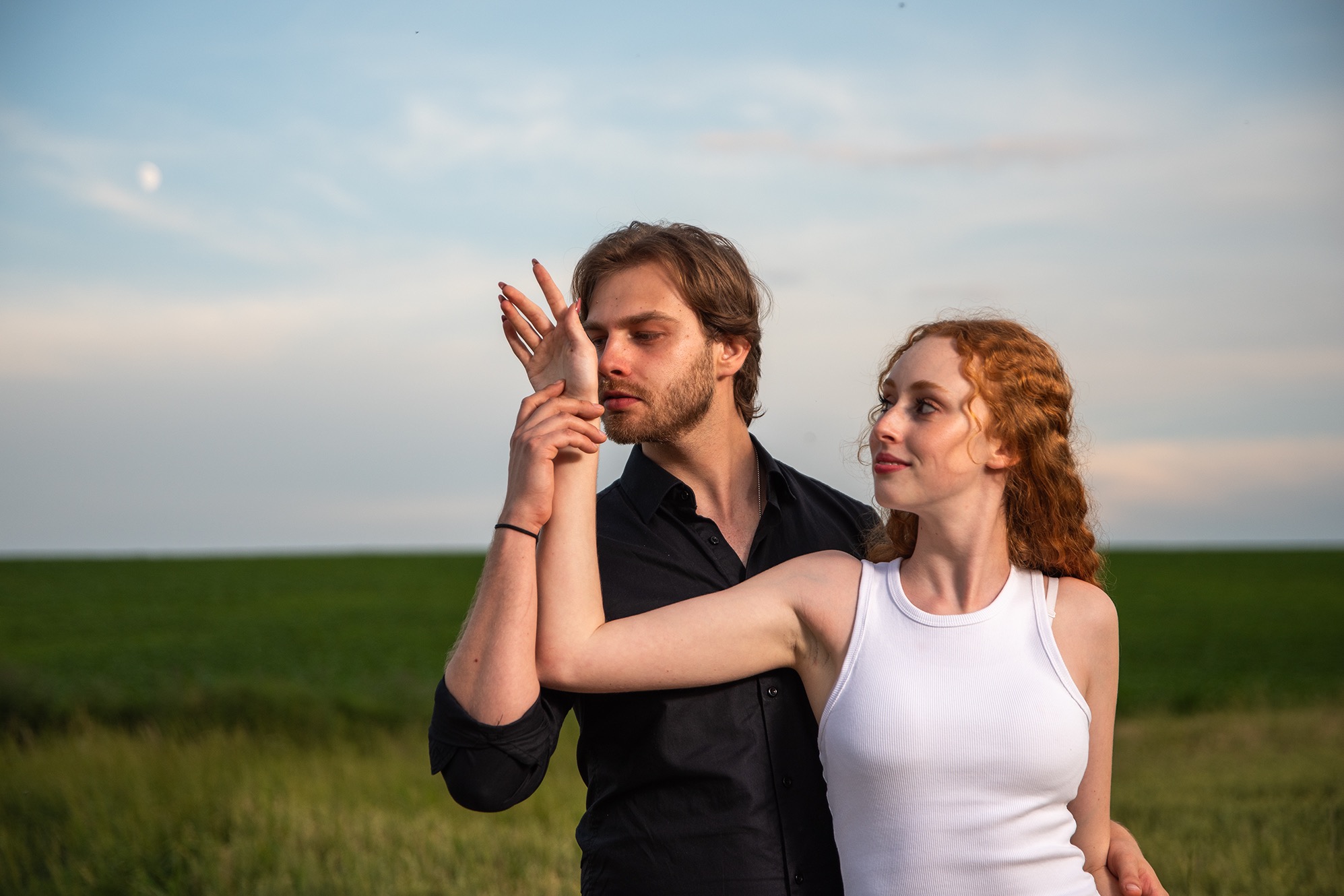 Romantyczna sesja narzeczeńska w plenerze: mężczyzna w czarnej koszuli trzyma dłoń rudej kobiety w białym topie na tle zielonego pola i błękitnego nieba.