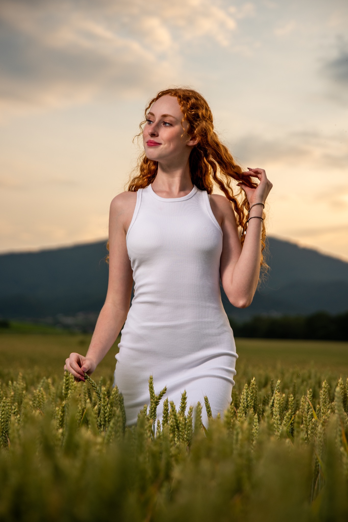 Radosna, rudowłosa kobieta w białej sukience, pozuje na tle pola zboża i zachodzącego słońca. Delikatny uśmiech, naturalny makijaż, subtelna biżuteria na nadgarstku.