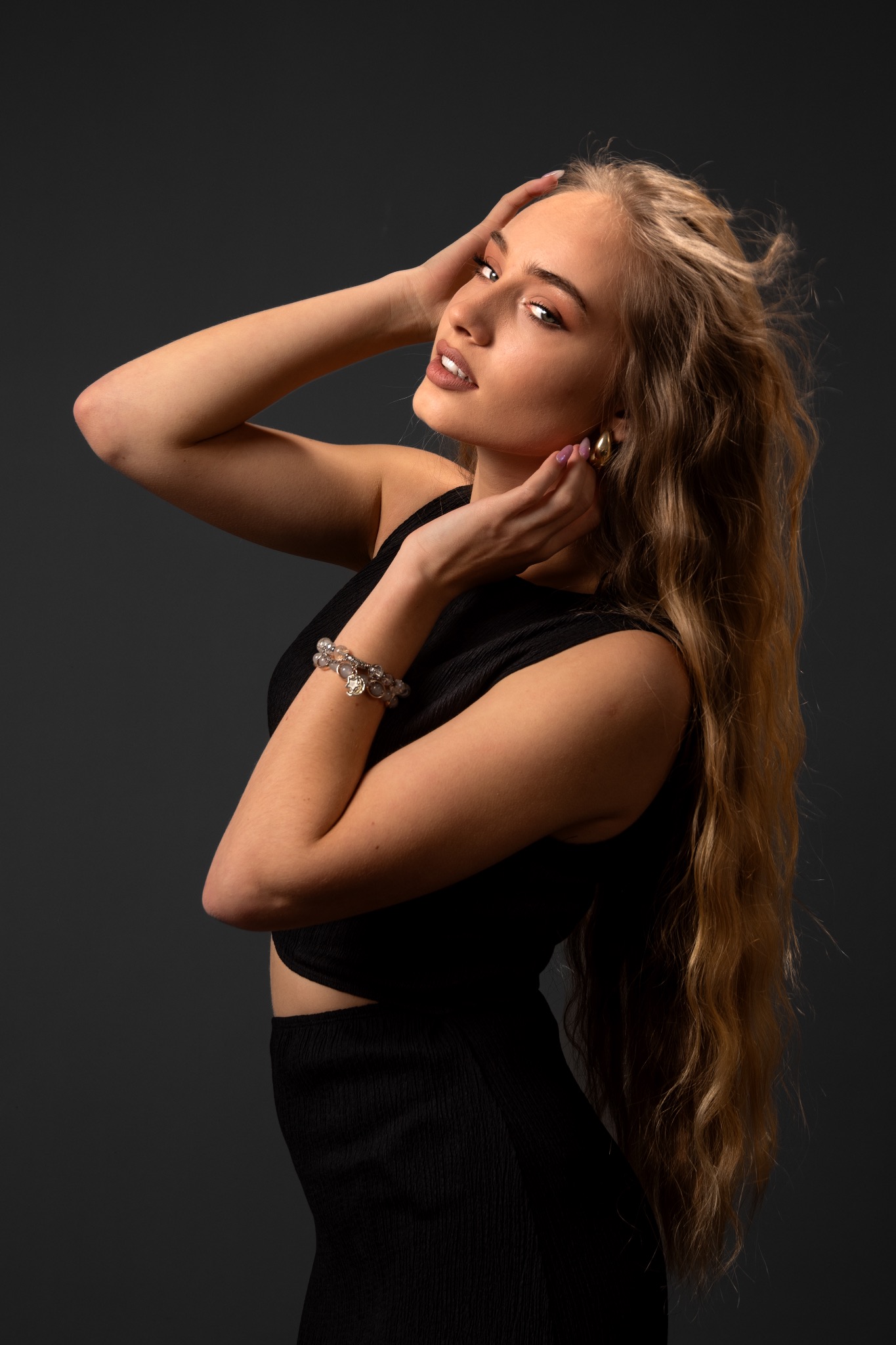 Portret kobiety w czarnej sukience, z rozwianymi blond włosami i biżuterią, na ciemnym tle. Sesja w Bielsko-Białej, profesjonalne oświetlenie.