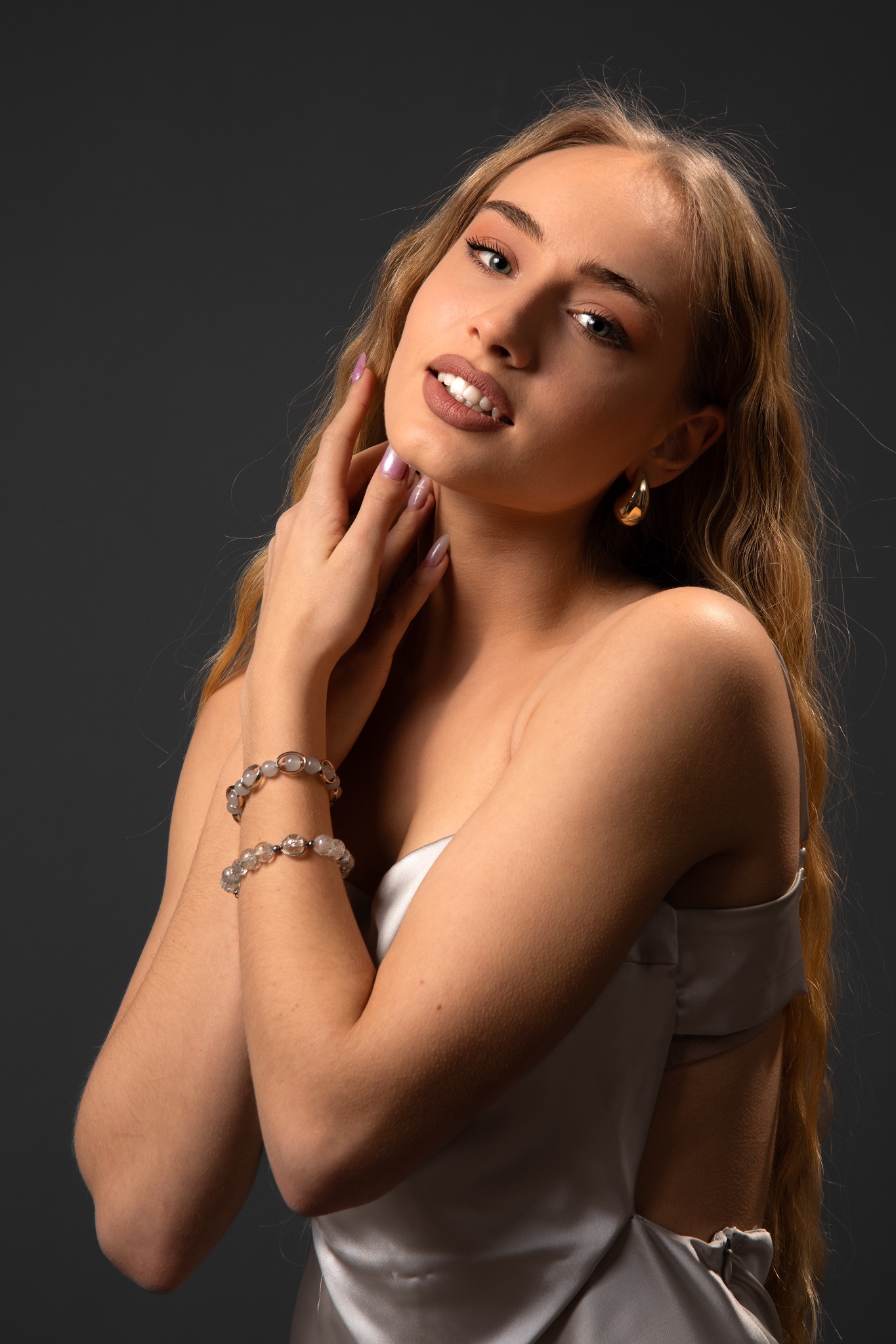 Portret kobiety z blond włosami, w satynowej sukience, z biżuterią na rękach i złotymi kolczykami, na ciemnoszarym tle. Uśmiech, makijaż, dłonie przy twarzy.