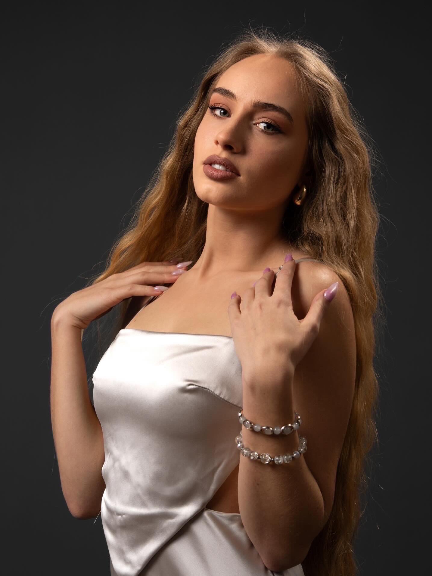 Portret kobiety w satynowej sukience na ciemnym tle. Widoczne detale: delikatny makijaż, biżuteria na ręce i złote kolczyki. Subtelne oświetlenie podkreśla urodę modelki.