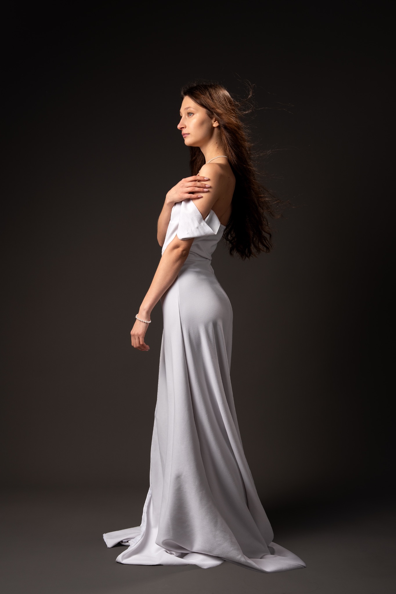 Elegancka kobieta w długiej, jasnej sukni pozuje bokiem na ciemnym tle. Delikatna biżuteria, rozwiane włosy i subtelny makijaż podkreślają jej naturalne piękno.