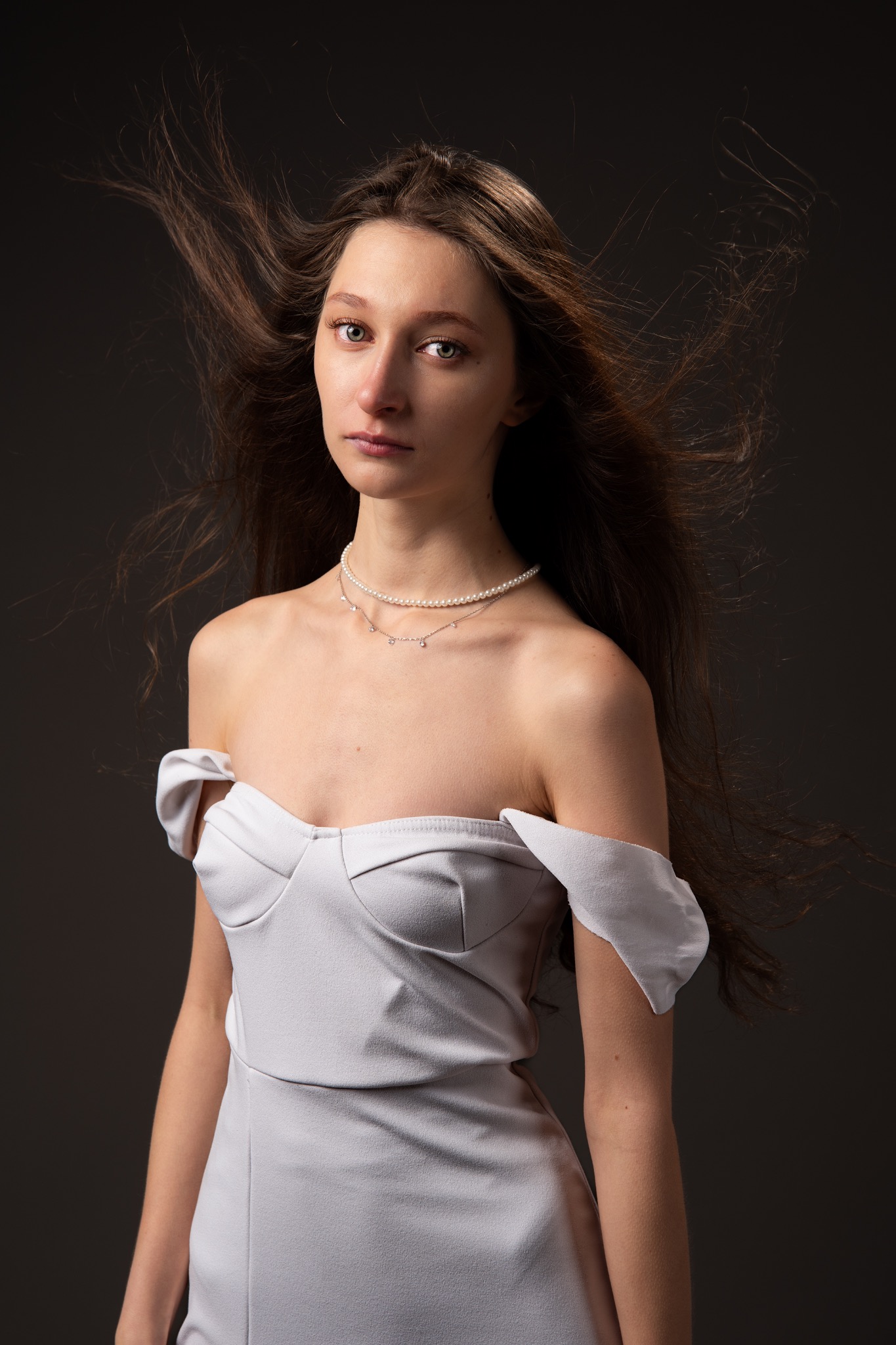 Portret kobiety w jasnej sukni z odkrytymi ramionami i perłowym naszyjnikiem, z rozwianymi włosami na ciemnym tle. Subtelne oświetlenie podkreśla rysy twarzy.