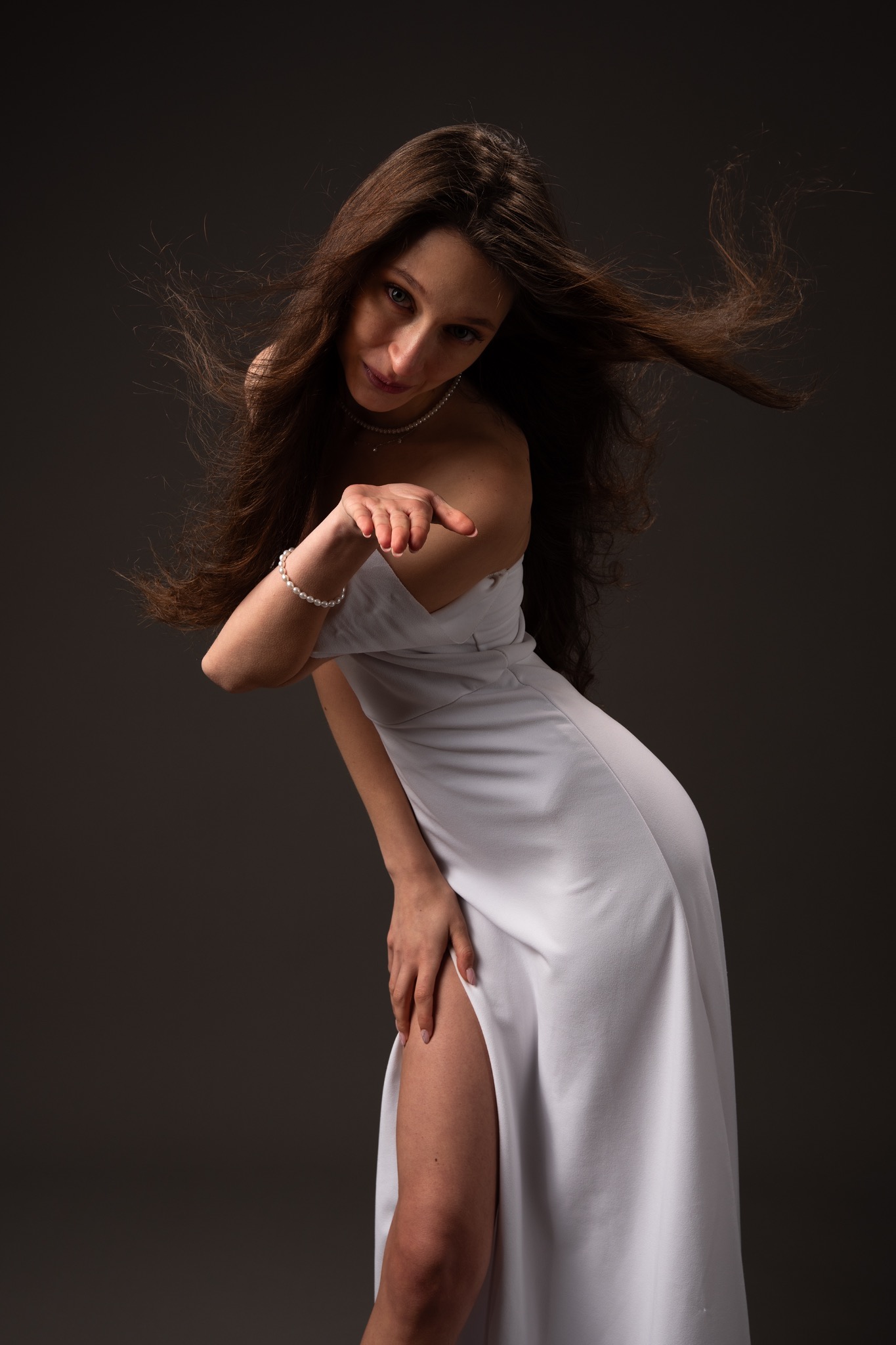 Kobieta w białej sukni z rozcięciem, z perłami na szyi i bransolecie, posyła całusa na ciemnym tle, włosy w ruchu.