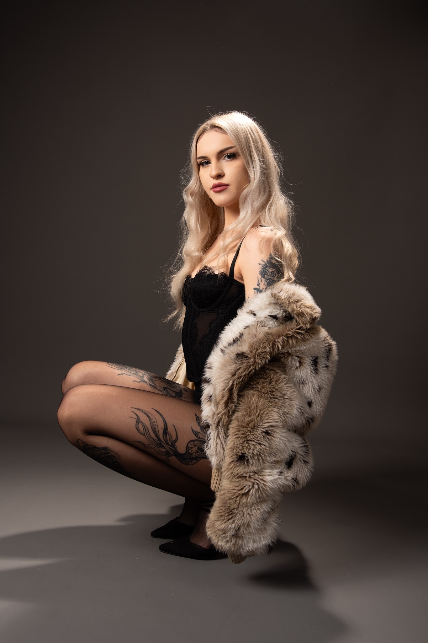 Kobieta w czarnym body i futrze pozuje w kucki na szarym tle. Widoczne tatuaże na ramieniu i nogach. Delikatny makijaż i blond włosy dodają elegancji.