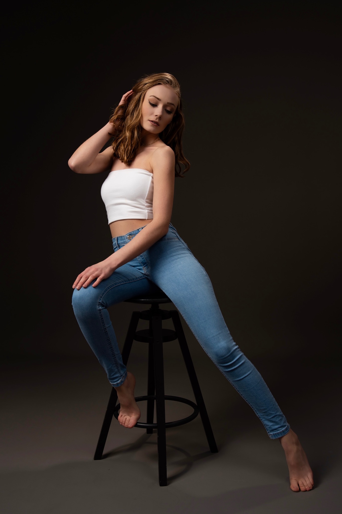 Portret kobiety siedzącej na stołku barowym, ubrana w jeansy i biały top, z delikatnym uśmiechem i dłonią we włosach, na ciemnym tle. Subtelne oświetlenie podkreśla jej urodę.
