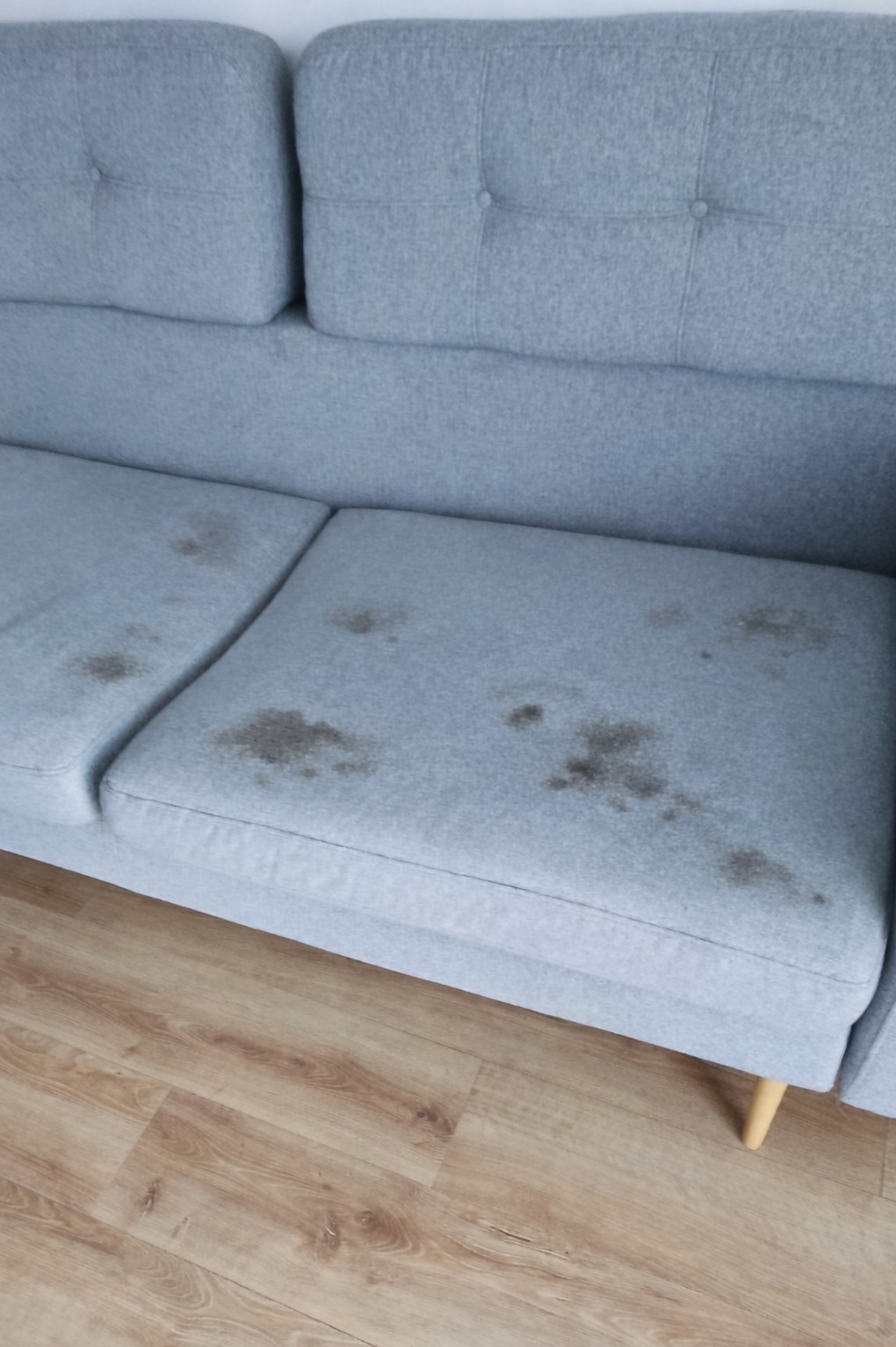 Szara kanapa z widocznymi plamami na siedzisku, stojąca na drewnianej podłodze. Widok z bliska, ukazujący zabrudzenia przed czyszczeniem.