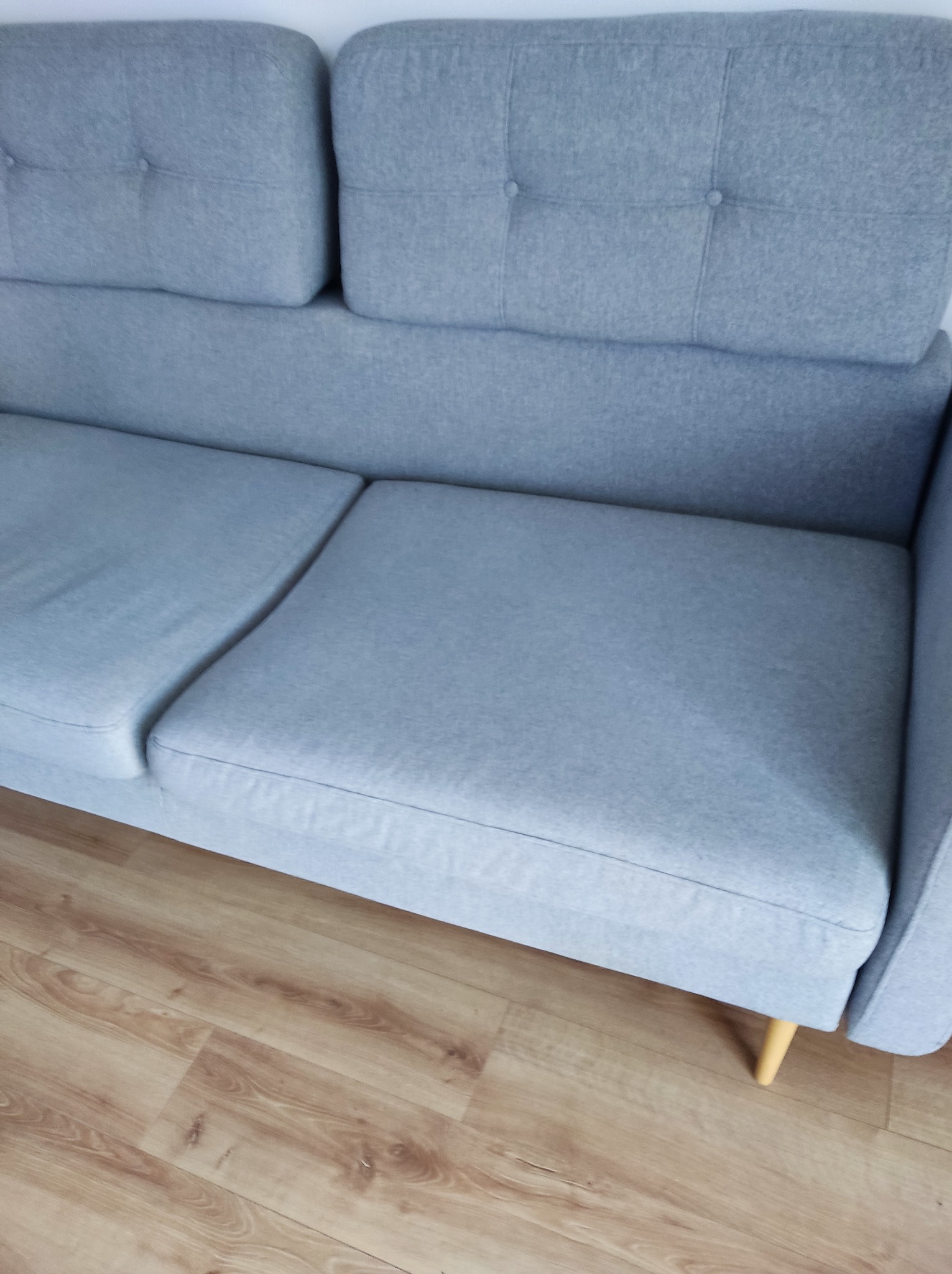 Jasnoszara kanapa z tapicerowanymi guzikami na oparciu, ustawiona na drewnianej podłodze. Widoczne nóżki kanapy. Ujęcie z góry, prezentujące czystą tapicerkę.