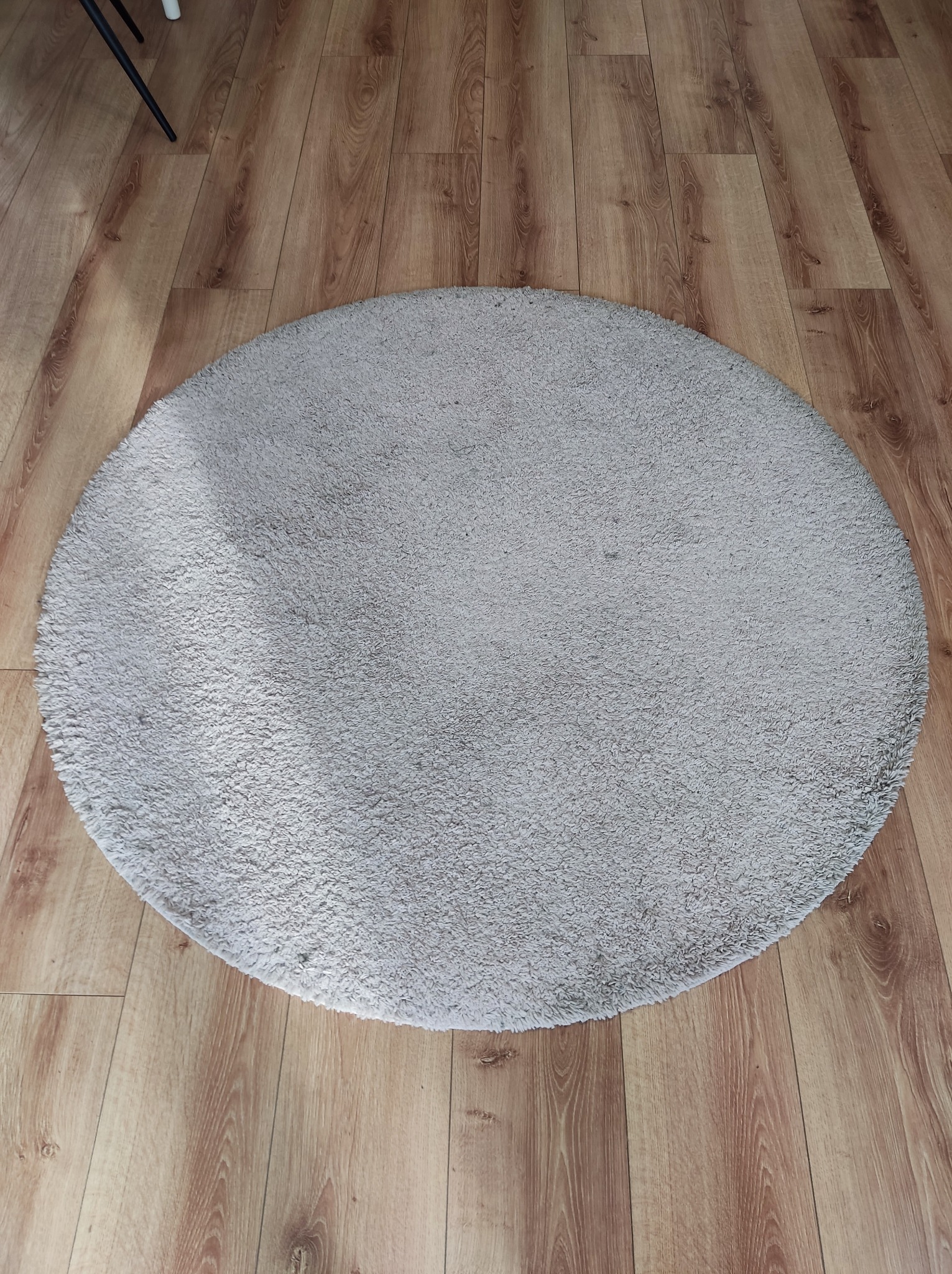 Okrągły, jasnoszary dywanik z krótkim włosiem leży na podłodze z paneli imitujących drewno. Widoczny fragment nogi krzesła w lewym górnym rogu.