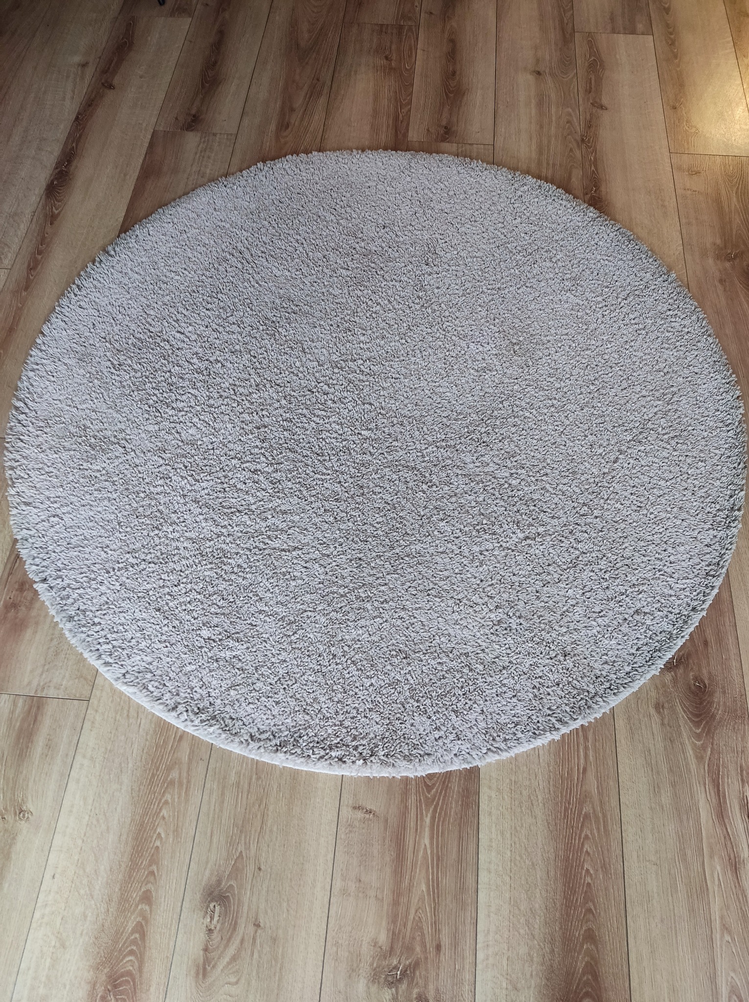 Okrągły, jasnoszary dywanik z krótkim włosiem leży na panelach podłogowych o wzorze drewna. Widoczne delikatne cieniowanie na dywanie i podłodze.