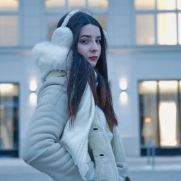 Kamil.photovibe - Portret kobiety w zimowej stylizacji na tle budynku z oknami, delikatny efekt padającego śniegu, stonowana kolorystyka, ciemne włosy, czerwone usta, futrzane nauszniki.