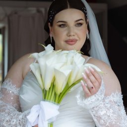 Kamil.photovibe - Panna młoda w białej sukni, z welonem i bukietem kalii, spogląda w bok. Delikatne zdobienia na włosach i koronkowe rękawiczki dodają elegancji. Subtelny makijaż podkreśla urodę.
