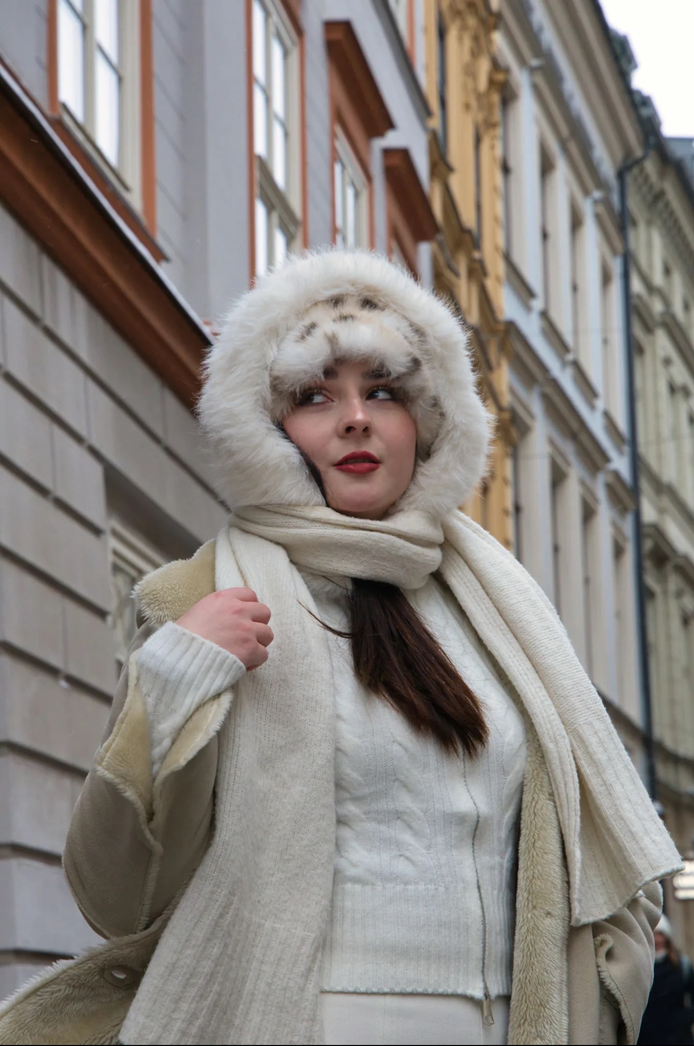Portret kobiety w zimowej stylizacji na tle kamienic. Biała futrzana czapka z kapturem i szal, jasny sweter. Ujęcie z dołu, podkreślające architekturę.