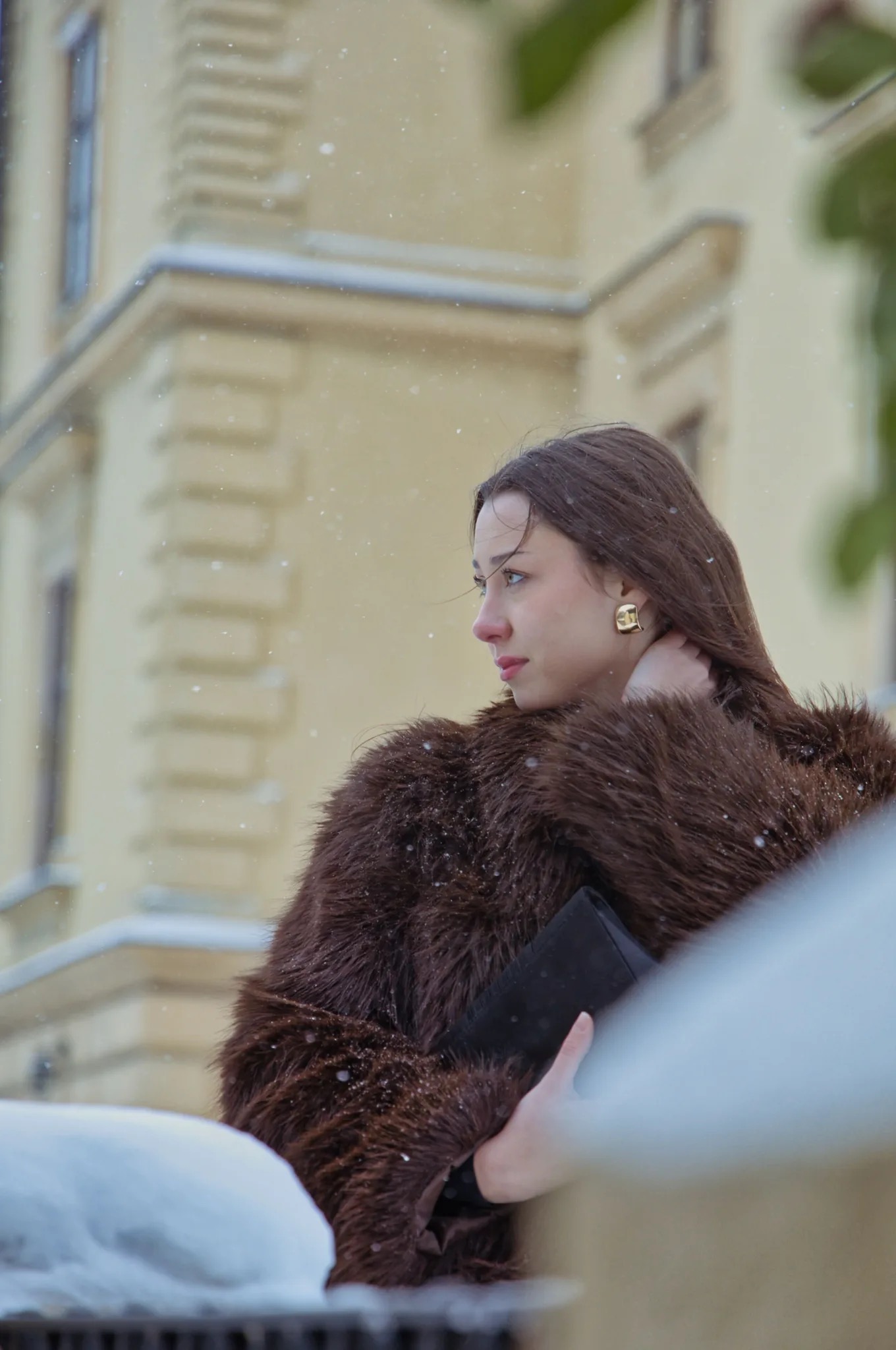 Portret kobiety w brązowym futrze na tle architektury w zimowej scenerii. Profil, śnieg, złote kolczyki, elegancki wygląd. Ujęcie z dołu, podkreślające urodę modelki.