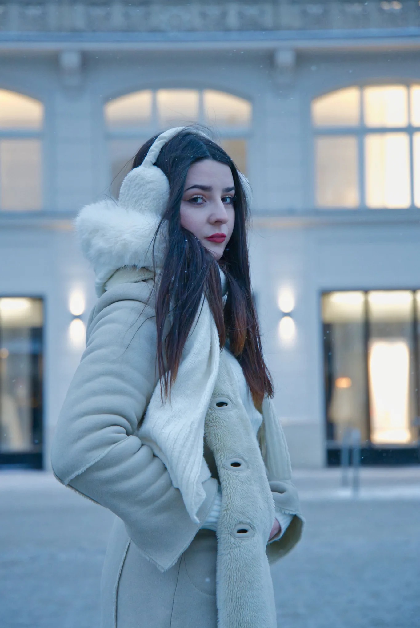Portret kobiety w zimowej stylizacji na tle budynku z oknami, delikatny efekt padającego śniegu, stonowana kolorystyka, ciemne włosy, czerwone usta, futrzane nauszniki.