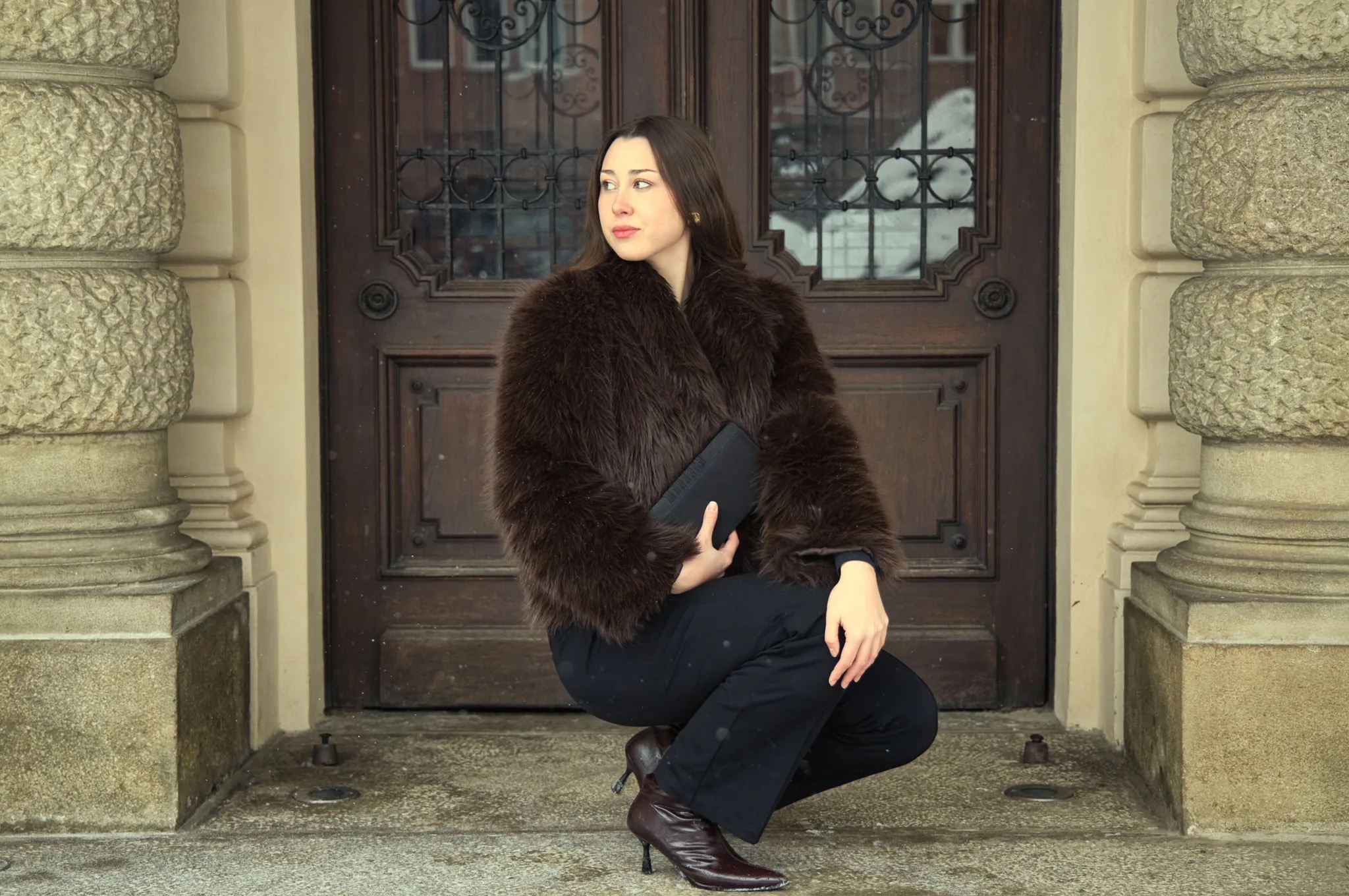 Kobieta w brązowym futrze kuca na tle zdobionych drzwi i kolumn. Ubrana w czarne spodnie i bordowe botki na obcasie, trzyma czarną kopertówkę.
